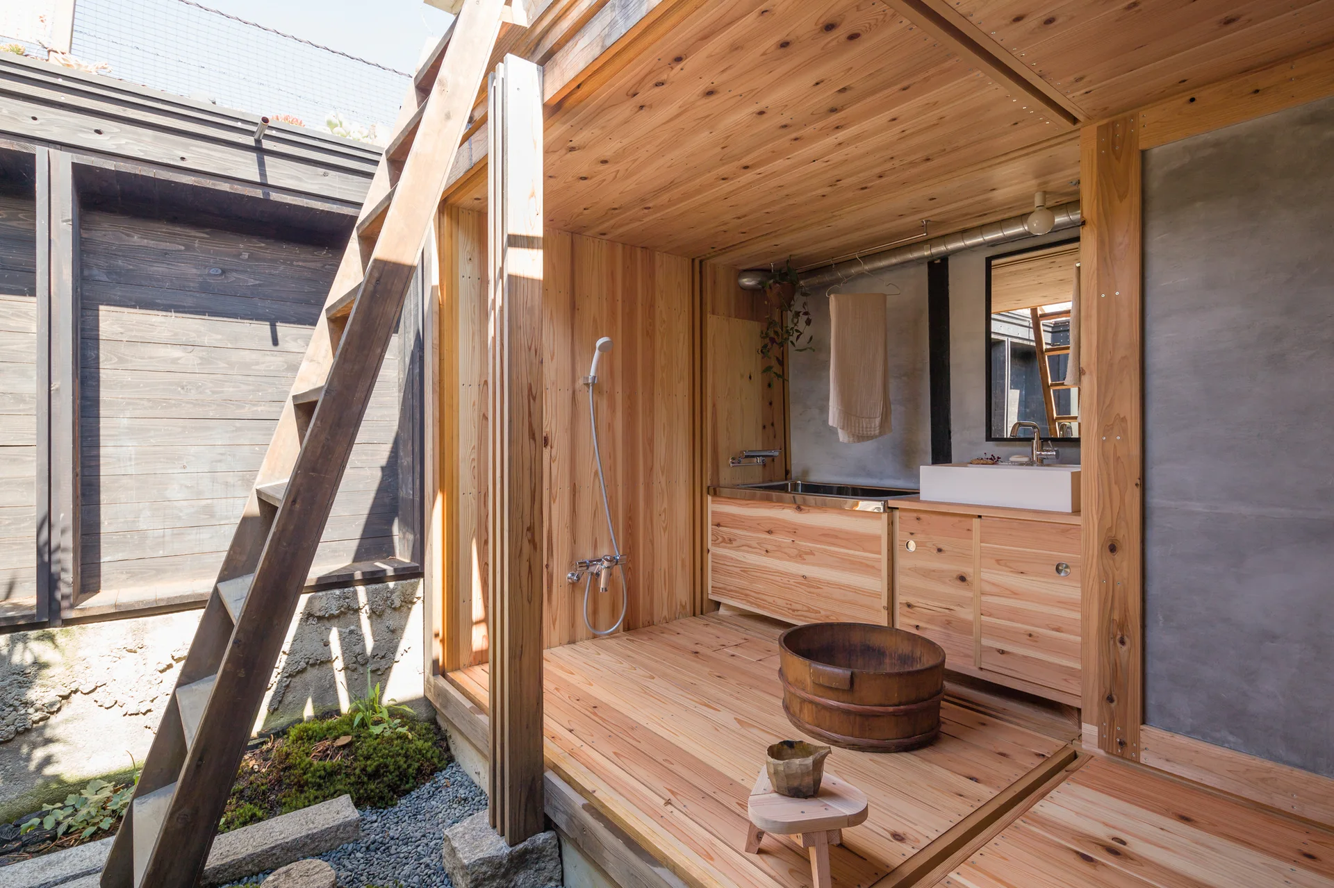 A minimalist Japanese style bathroom features cedar wood walls, a concrete panel, a wooden vanity with a white basin, and a traditional wash bucket.