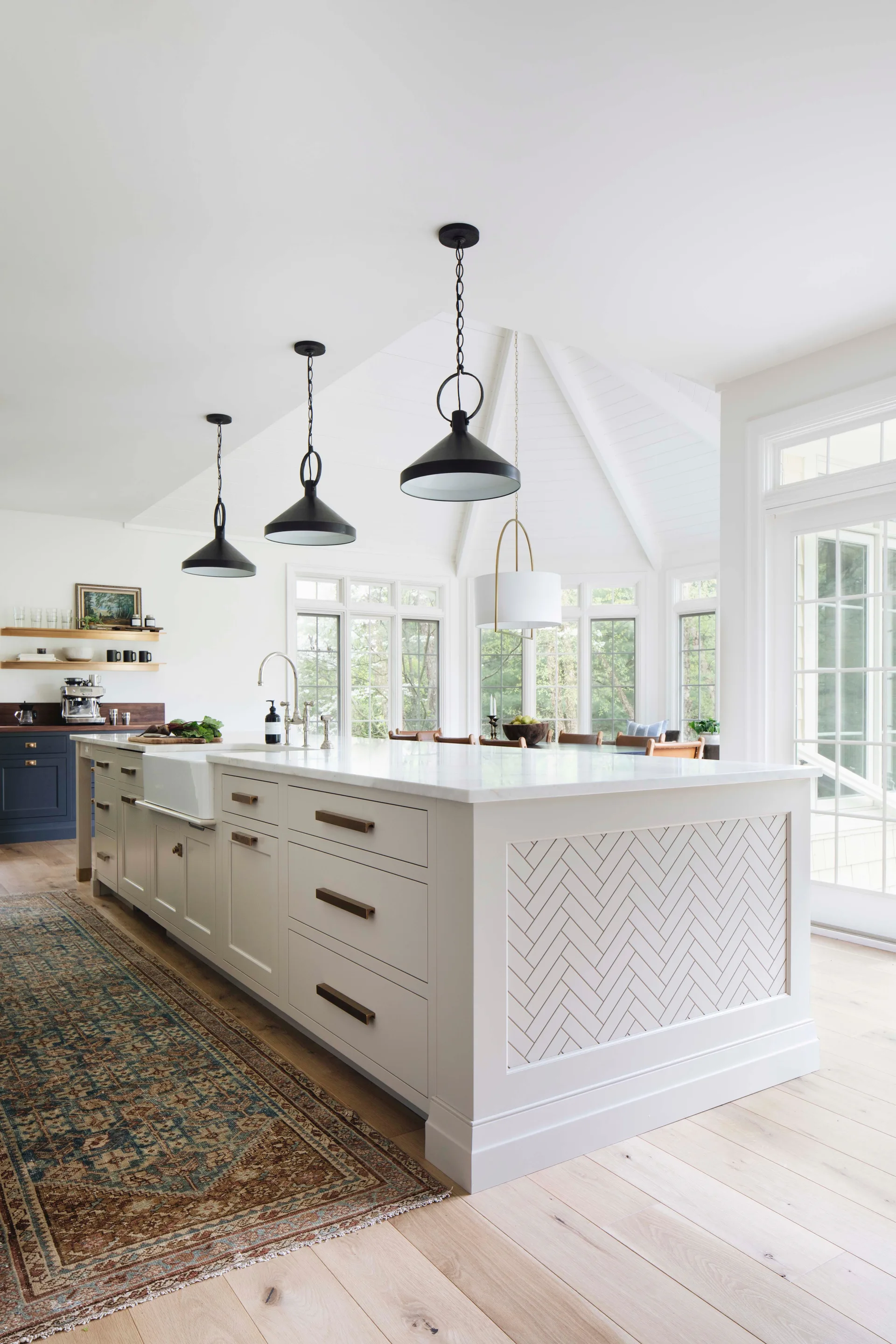 A spacious white kitchen featuring a large central island with a herringbone pattern side panel, black pendant lights, and a patterned runner rug.
