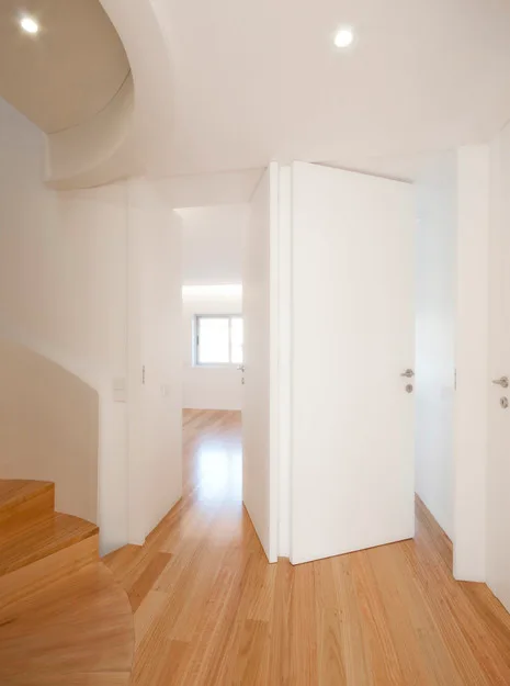 A bright white hallway features light wood plank flooring, several flush-panel white doors, and a curved wooden staircase on the left.