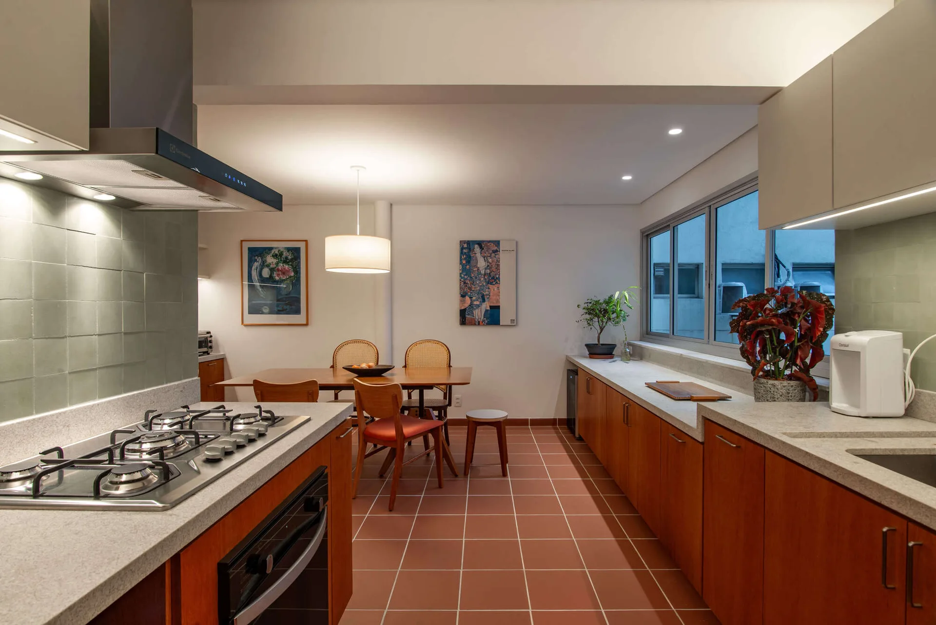 An open-plan kitchen features warm wood cabinetry, a sage green tiled backsplash, and terra-cotta floor tiles next to a wooden dining set.