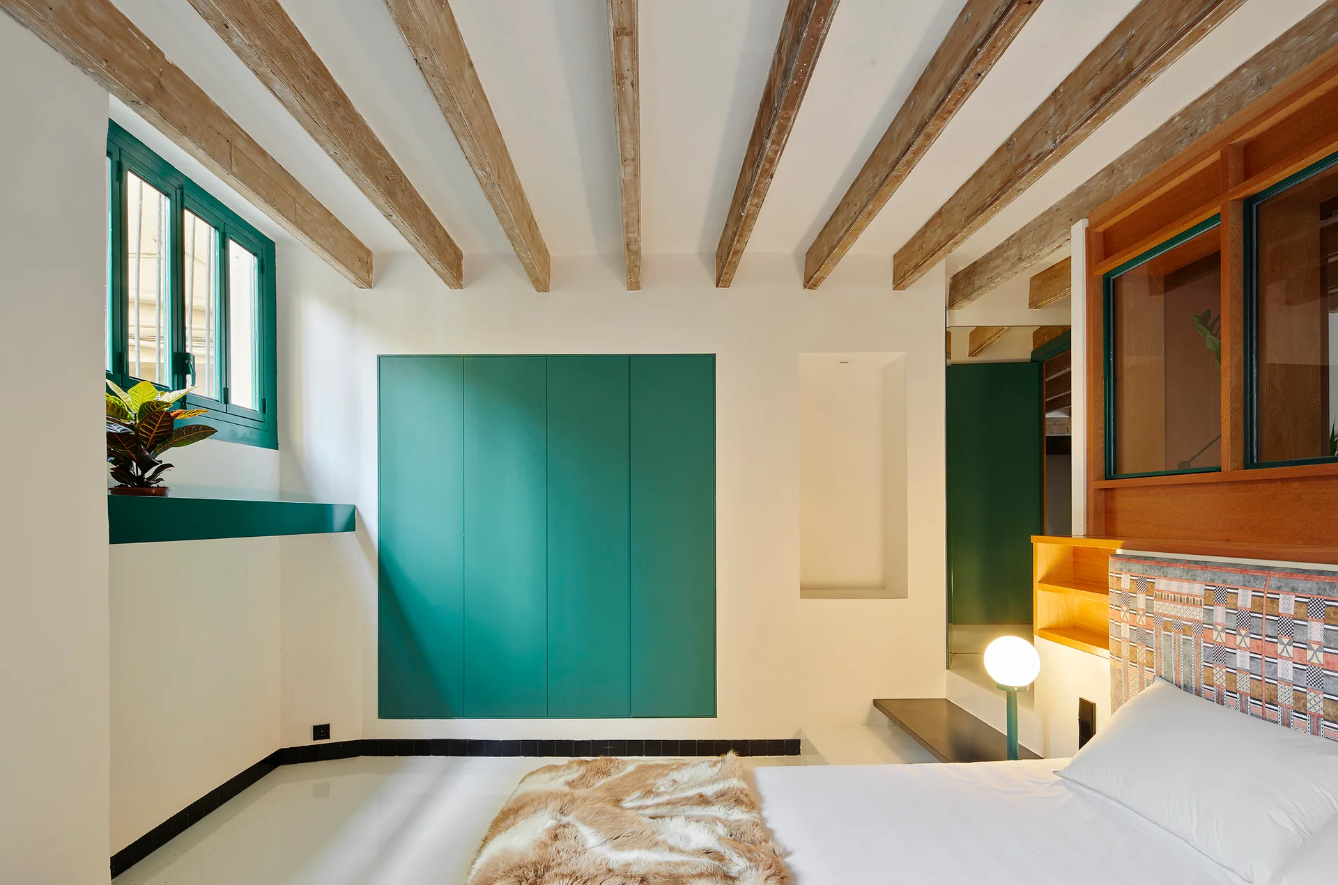A modern bedroom with white walls, exposed wood ceiling beams, a bed with a patterned headboard, and a large green built-in cabinet.