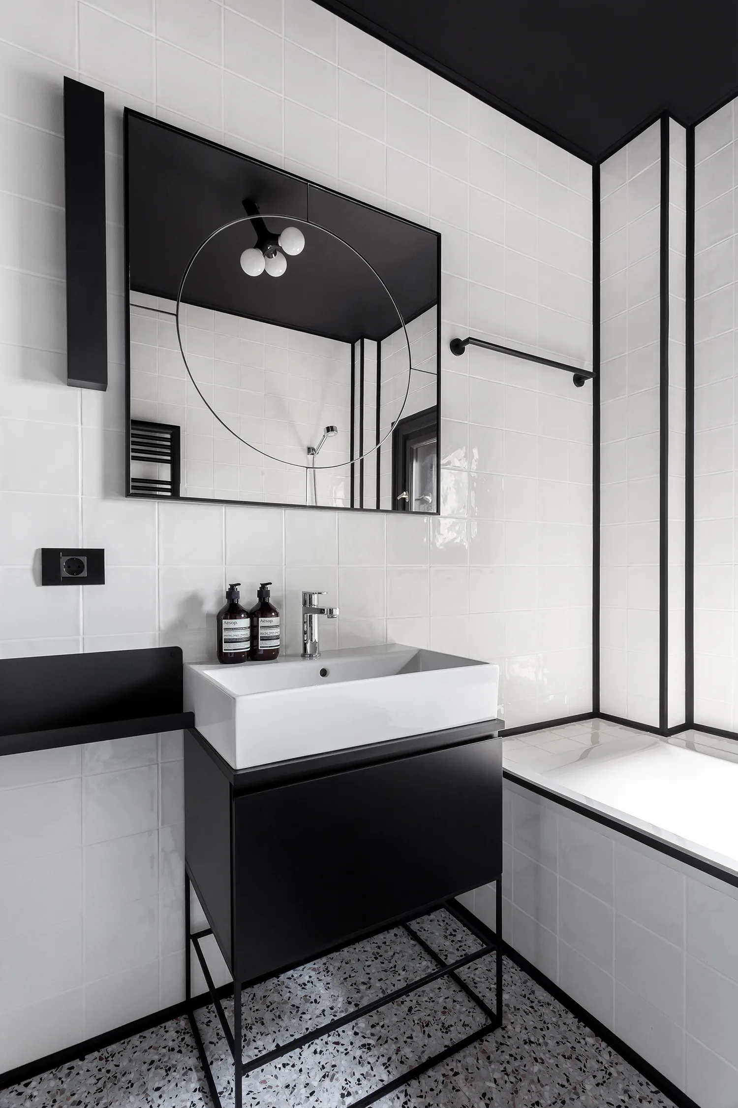 A modern bathroom featuring white square wall tiles with black trim, a black metal vanity with a white vessel sink, and terrazzo tile floors.