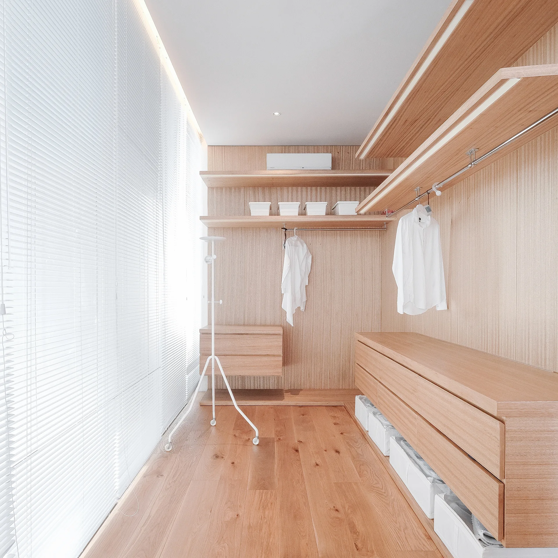 A minimalist walk-in closet features light wood wardrobes, vertical paneled walls, wood plank flooring, and a large window covered in white slatted blinds.
