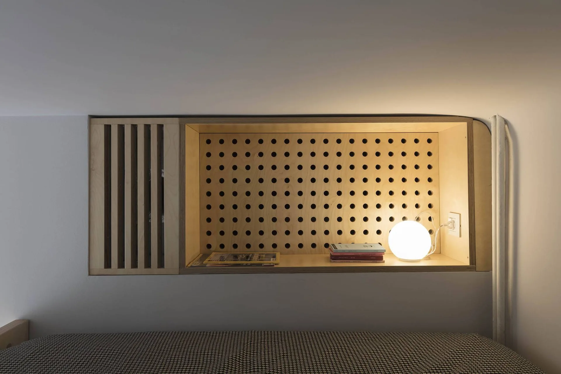 A custom recessed plywood wall niche in a bedroom features a pegboard backing, vertical wood slats, and a glowing globe table lamp.