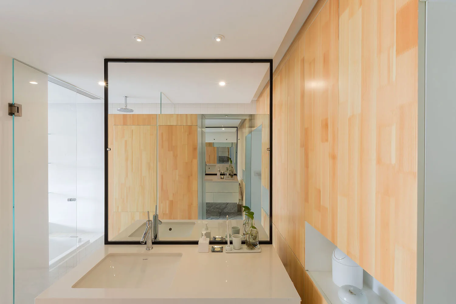 A minimalist bathroom features a white vanity sink, a large black-framed mirror, and light wood wall paneling.