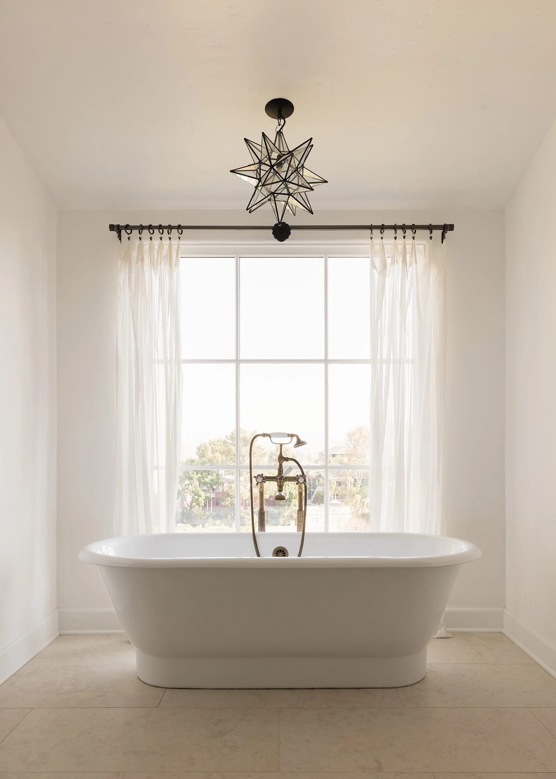 A white freestanding bathtub sits on a beige tiled floor in front of a window with sheer curtains beneath a glass star pendant light.