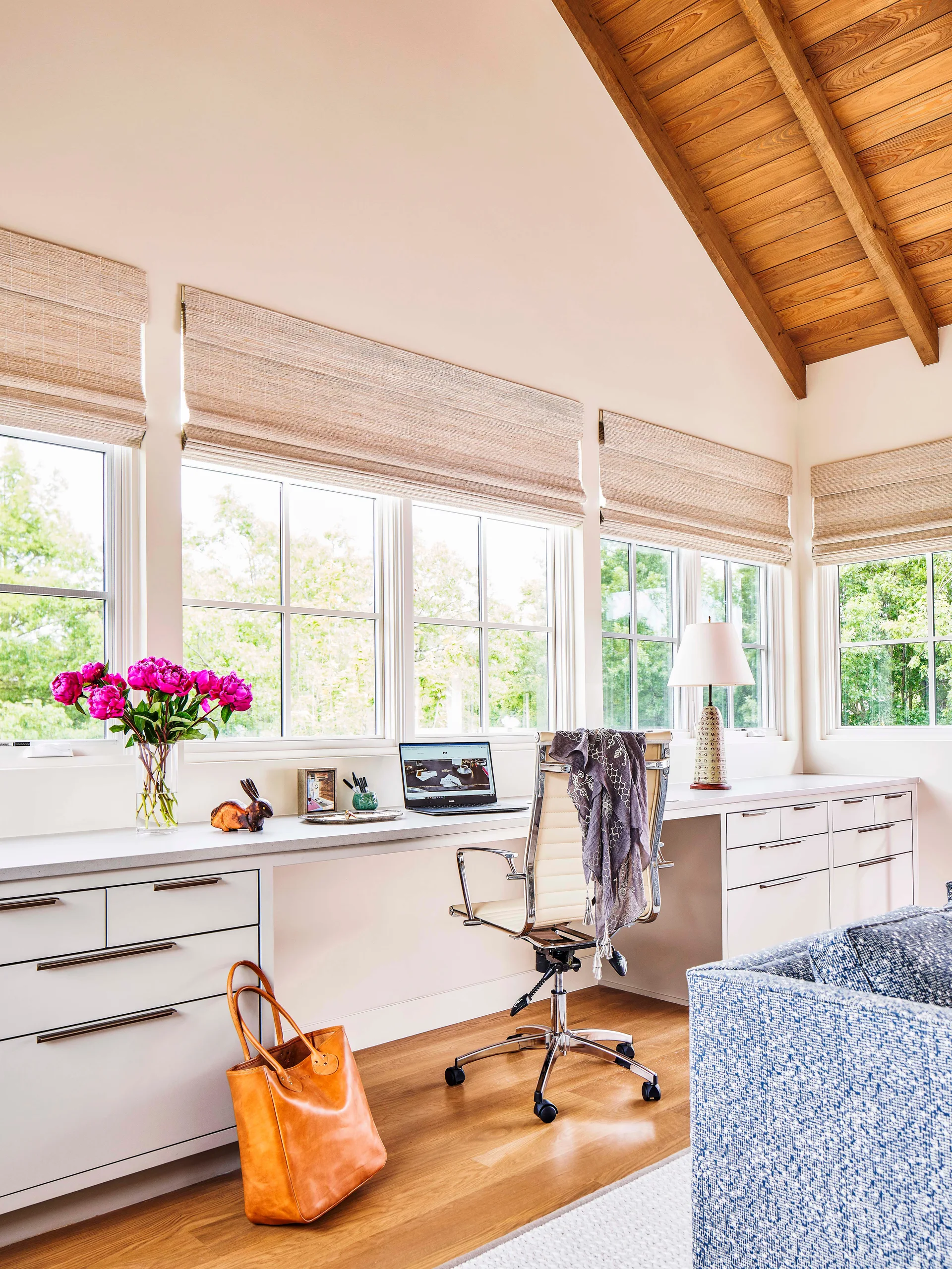 A bright home office features a long white built-in desk under four windows with woven roman shades below a vaulted wood beam ceiling.