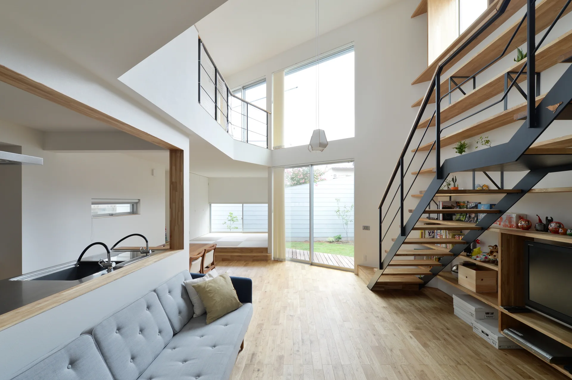 A bright open-concept living room featuring a grey sofa, light wood flooring, a modern floating staircase, and built-in shelving.