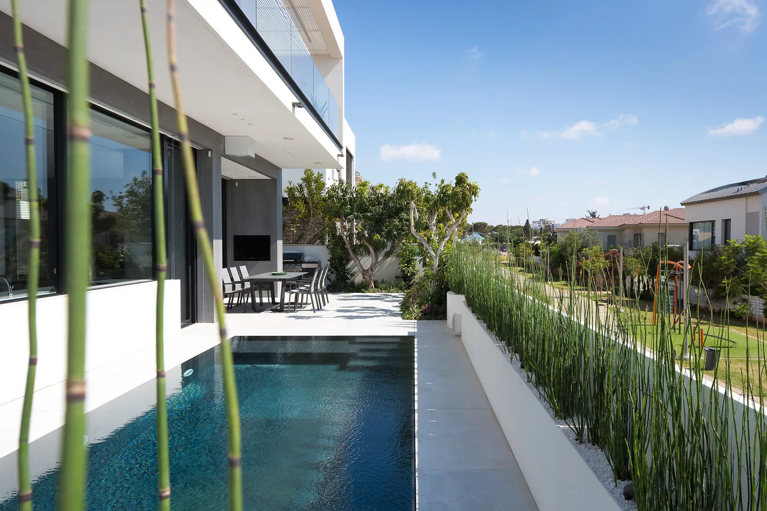 A rectangular swimming pool sits next to a paved patio featuring an outdoor dining set and a long white planter filled with tall green reeds.
