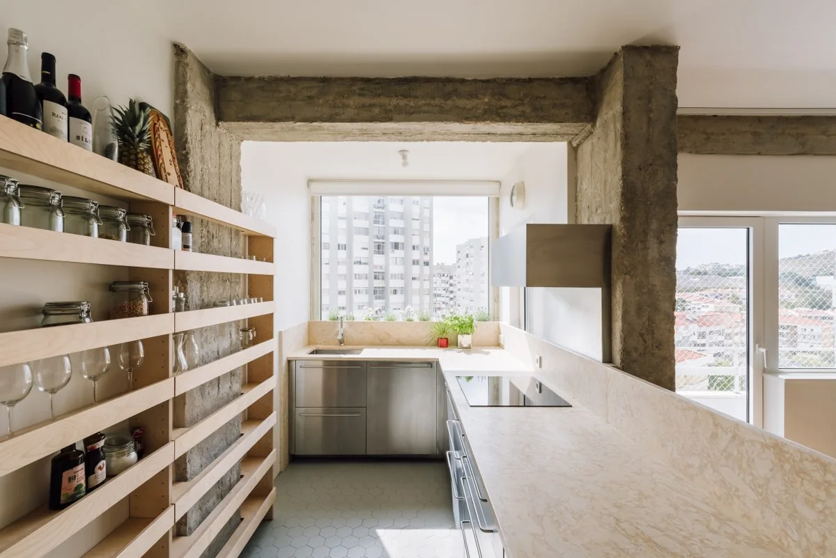 A modern kitchen featuring exposed concrete pillars, stainless steel cabinets, a light stone countertop, and custom open wooden shelving.
