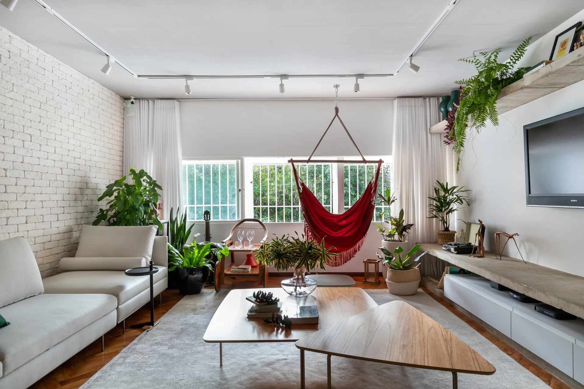 A bright living room features a white sectional sofa, nested wooden coffee tables, a red hanging hammock, and a white exposed brick wall.