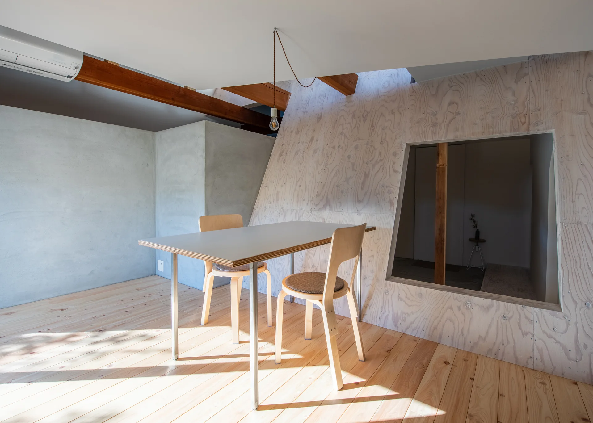 A minimalist dining area featuring a light wood table and chairs set against a slanted plywood paneled wall with a large geometric cutout opening.