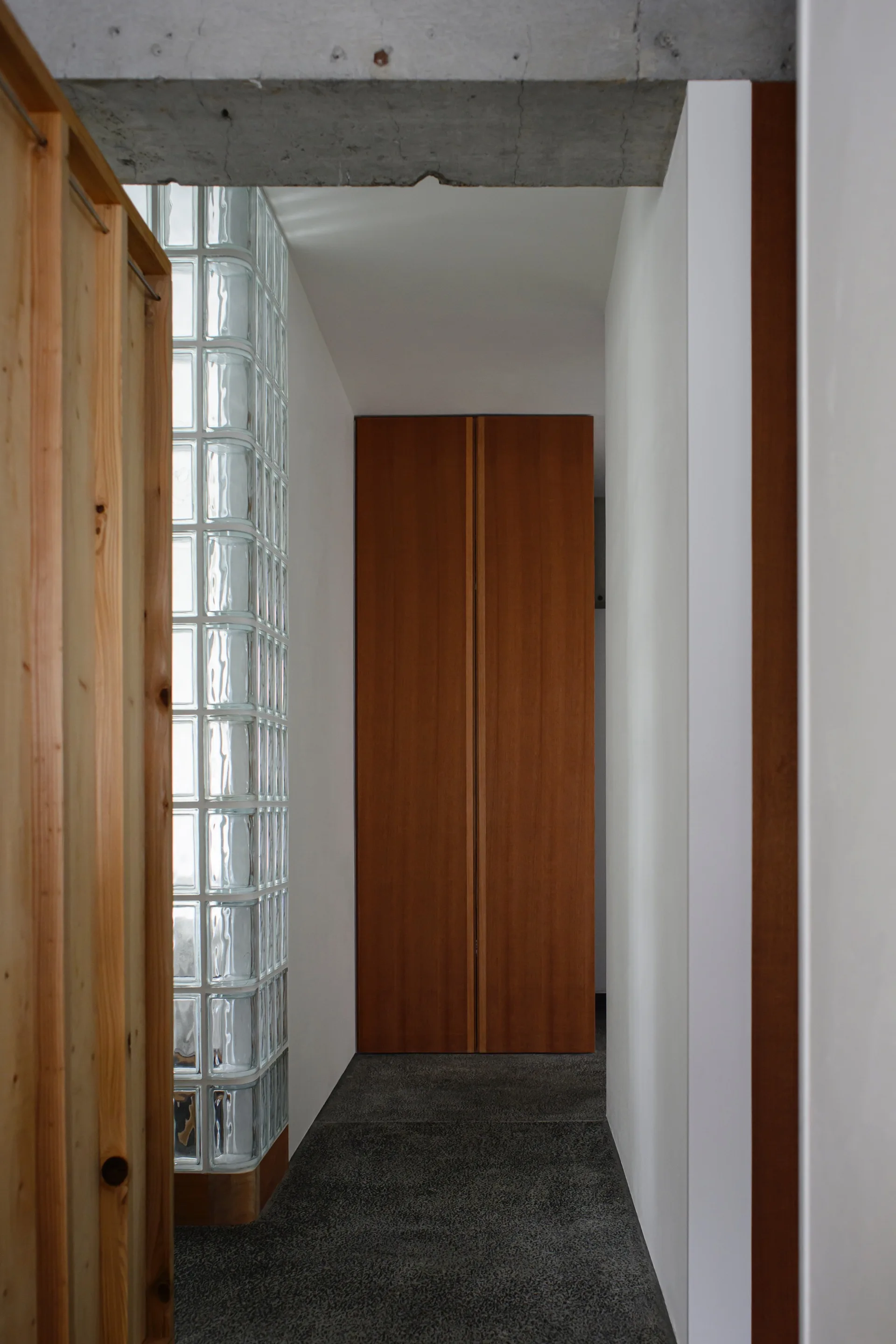 A narrow minimalist hallway features a glass block partition wall, an exposed concrete beam overhead, and a tall wood door at the end.
