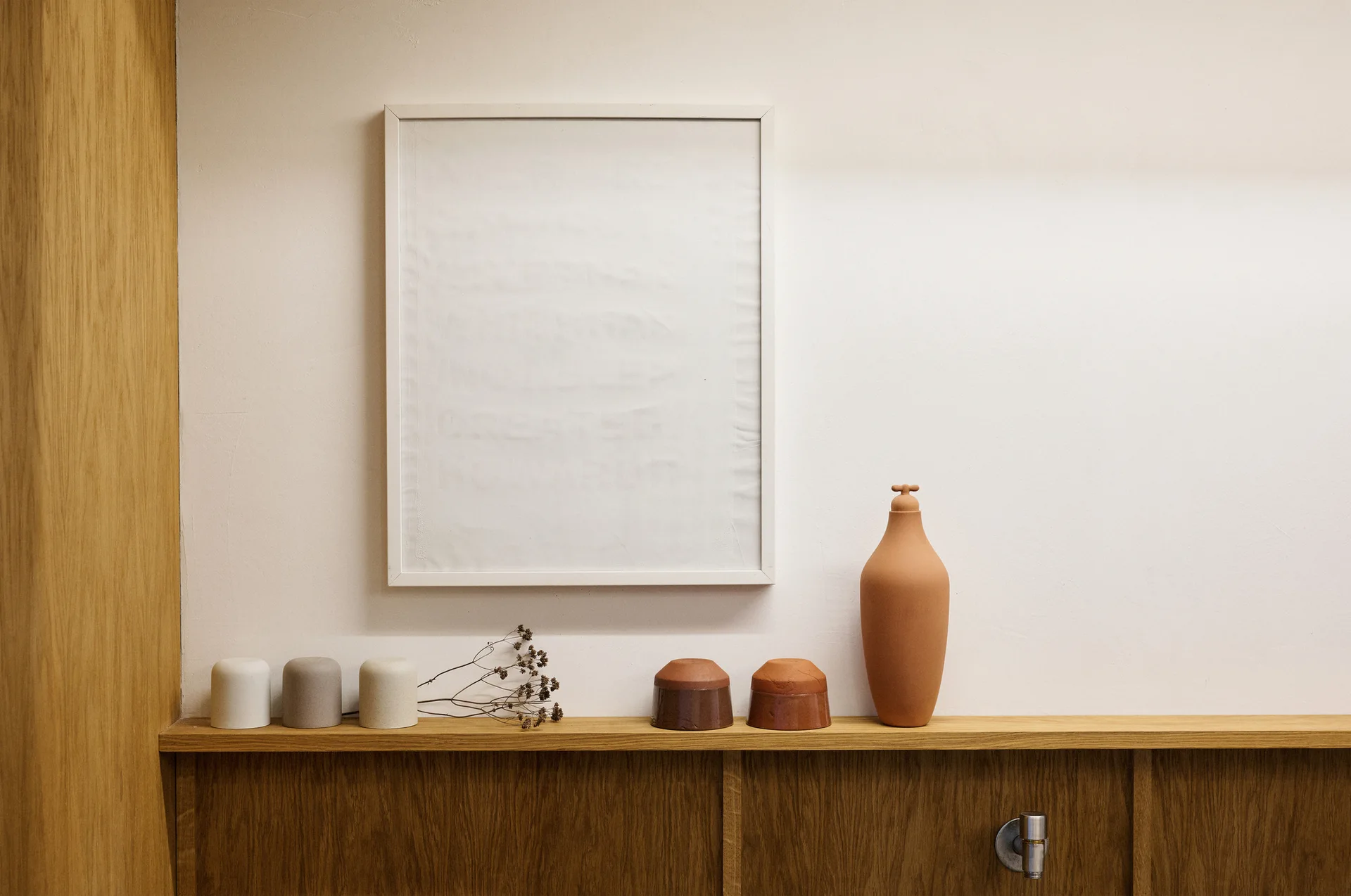Several earthy ceramic vessels and a single dried branch sit on a wood shelf below a white picture frame.