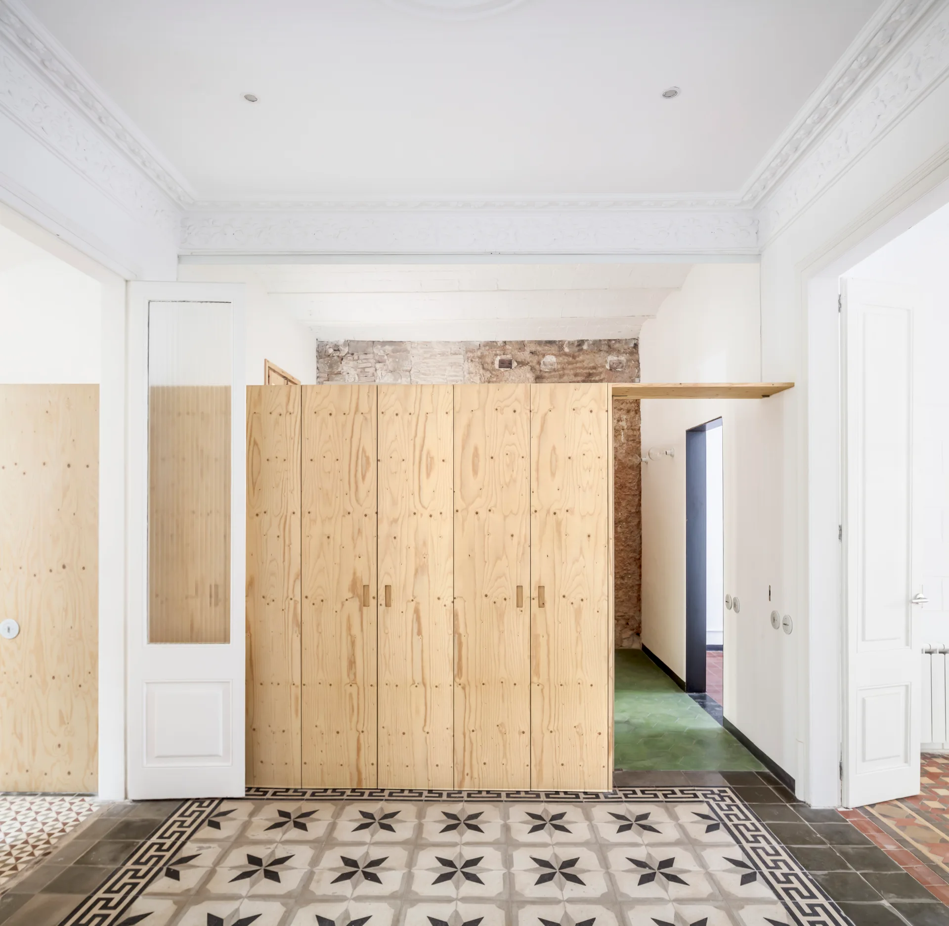 A central plywood partition stands in a bright white entryway featuring intricate patterned tile floors and ornate plaster ceiling molding.