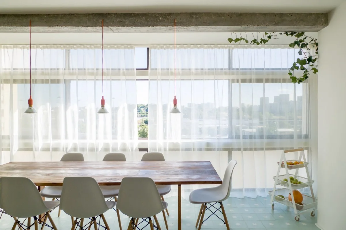 A wooden dining table with white shell chairs sits on a light blue tiled floor beneath three red cord pendant lights and an exposed concrete beam.