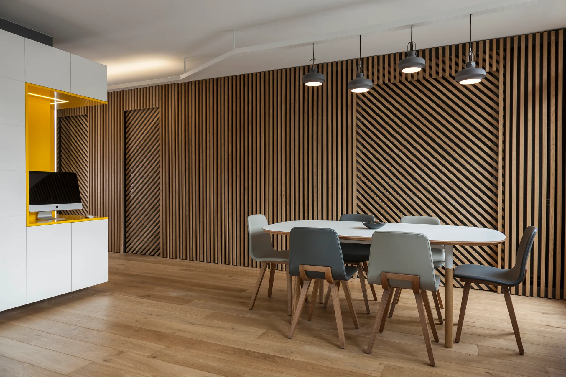 A modern dining room features a white oval table, grey chairs, four pendant lights, and a full wall of vertical and diagonal wood slats.
