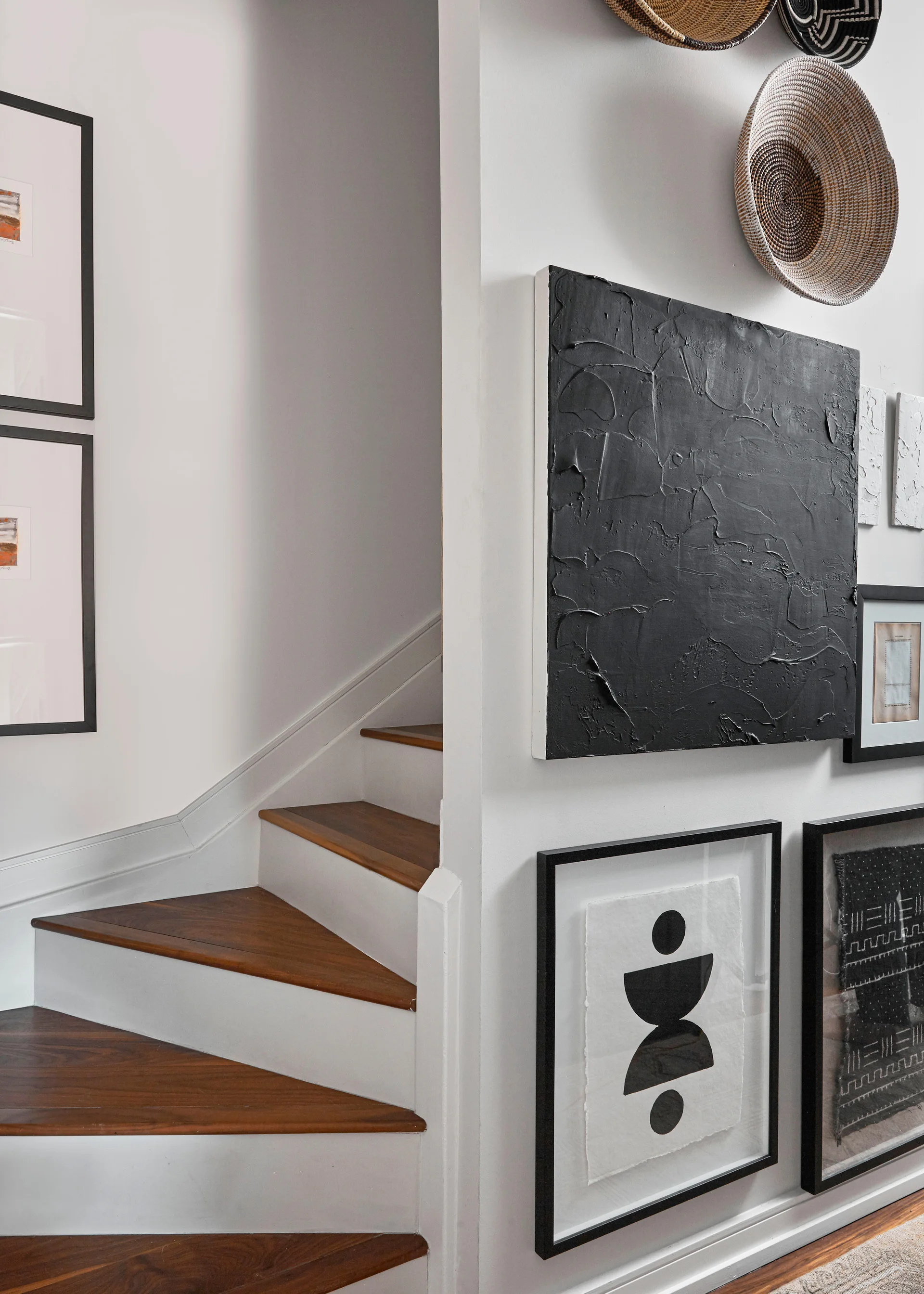 A modern staircase with wood treads next to a gallery wall showcasing textured black art, framed prints, and woven baskets.