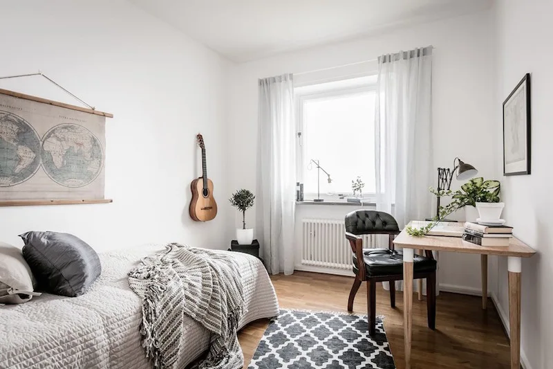 A small white bedroom features a single bed with textured grey bedding, a wooden writing desk with a black leather chair, and a guitar hanging on the wall.