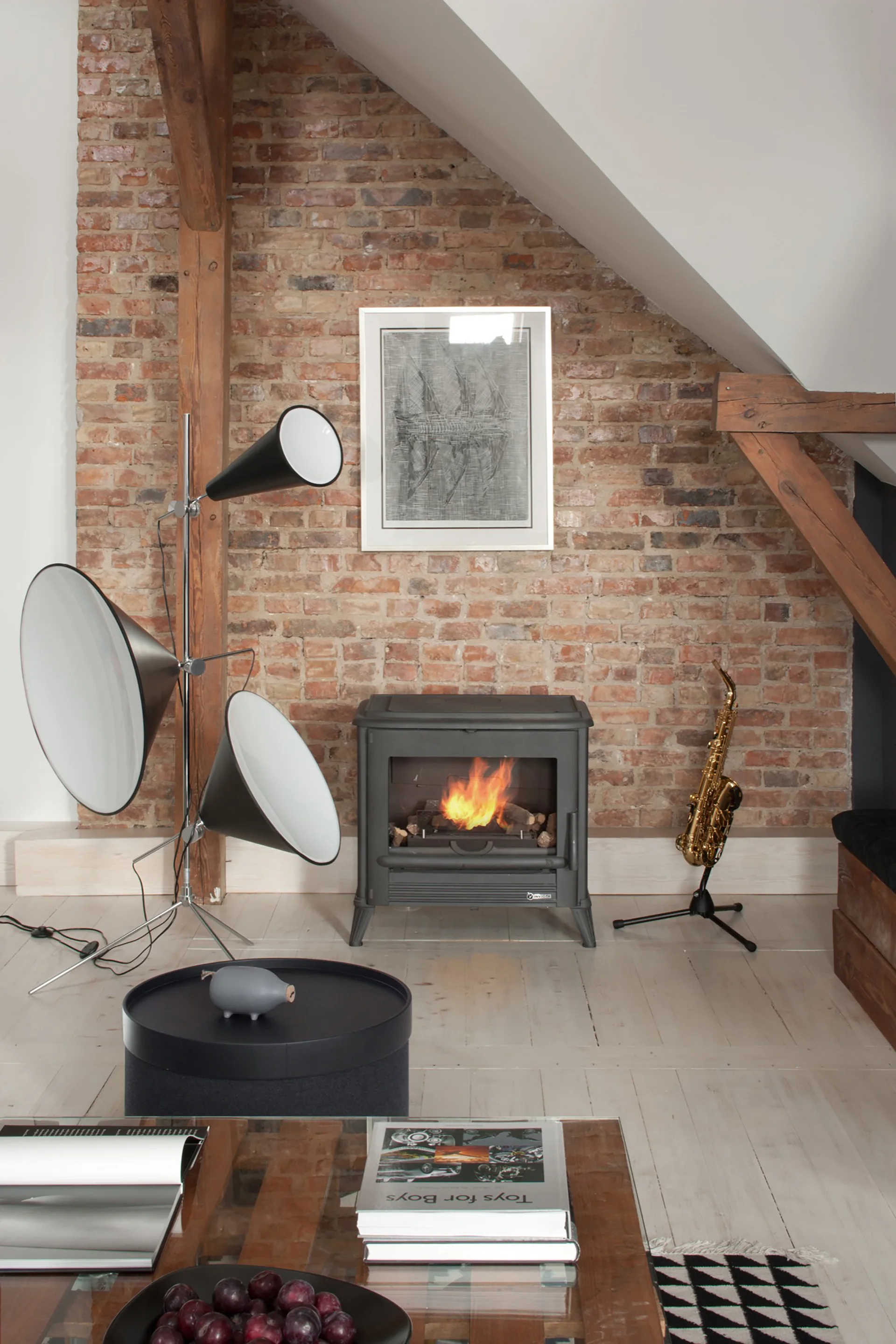 A black cast iron wood stove burns warmly against an exposed brick wall in an attic living room with white floors and timber beams.