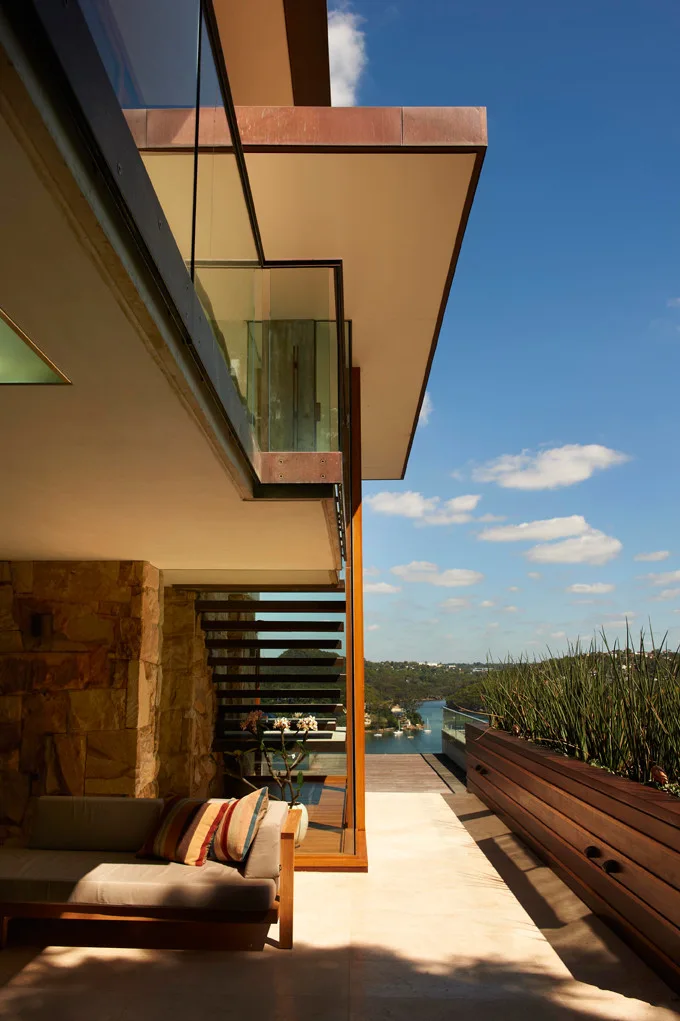 A modern outdoor terrace features a covered lounge area with a stone wall, an open-riser staircase, and a wood planter overlooking the water.
