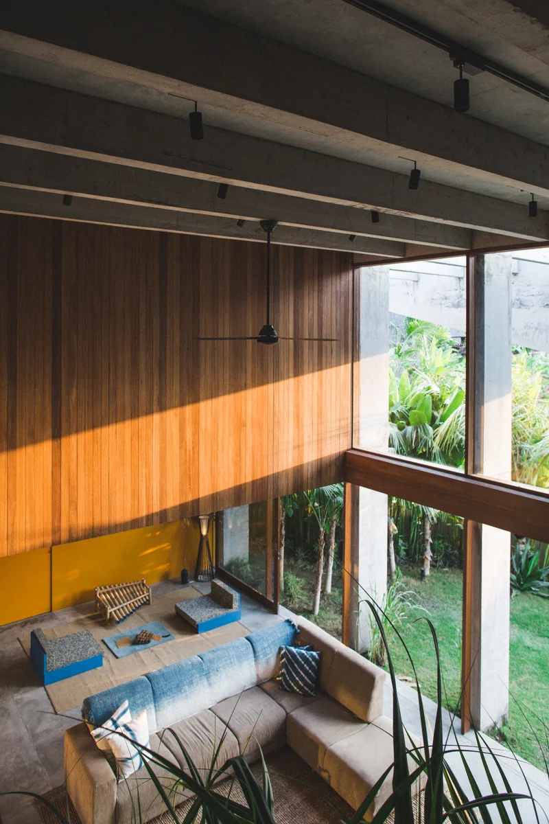 A double-height living room features a tan sectional sofa, vertical wood-paneled walls, exposed concrete ceiling beams, and large windows facing a garden.