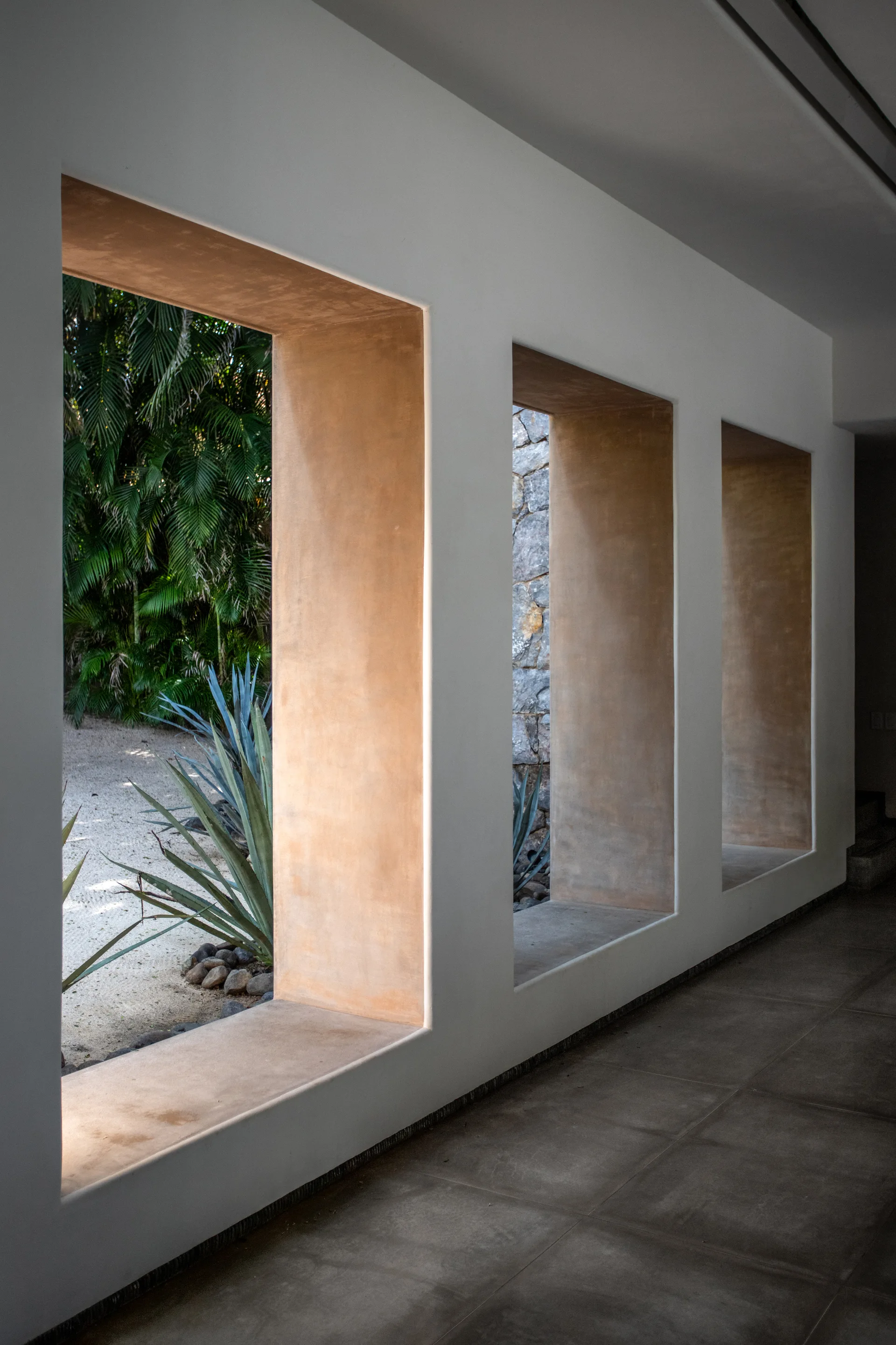 A minimalist hallway features three deep rectangular wall openings framed in warm tan plaster, offering views of a gravel garden with agave plants.