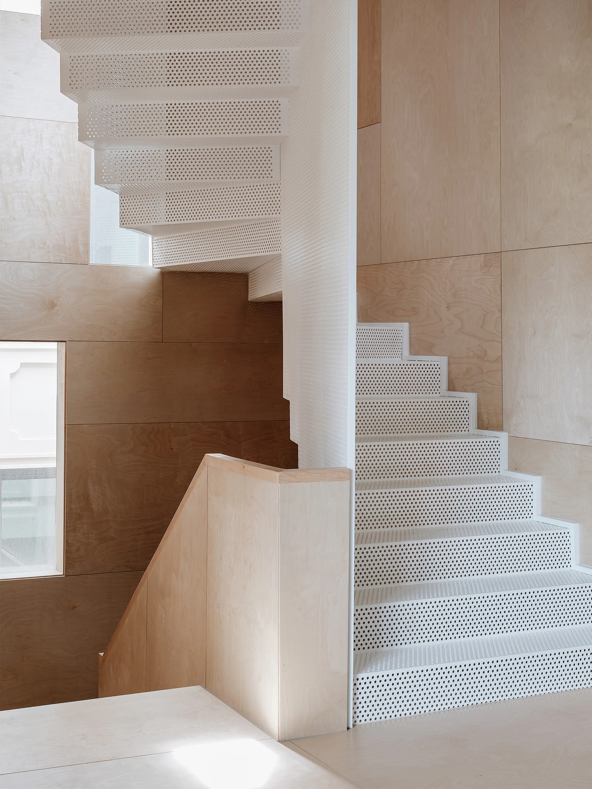 A white perforated metal spiral staircase winds upward against walls covered in light-toned plywood panels with recessed windows.