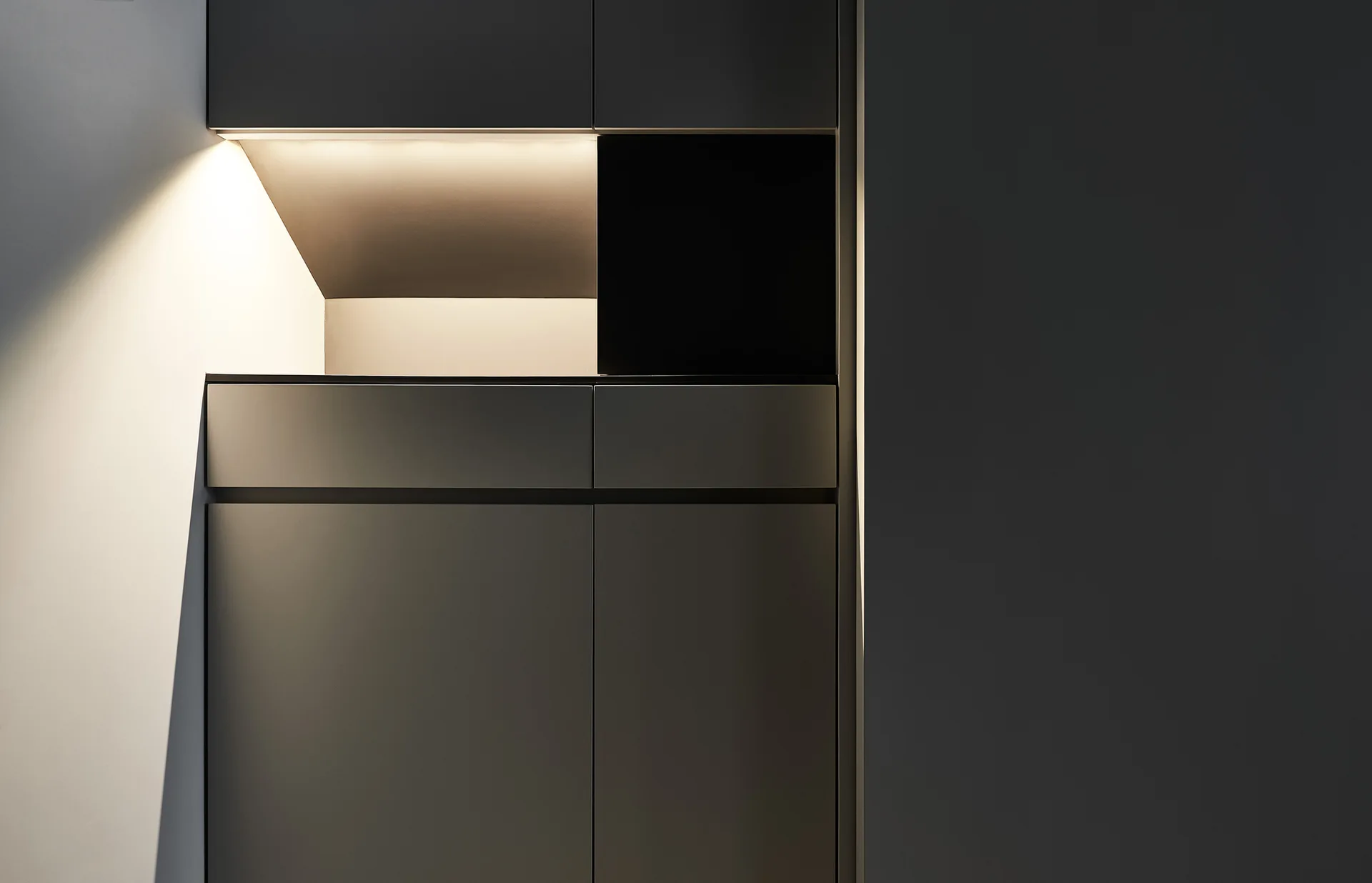 Minimalist charcoal gray kitchen cabinets feature a handleless design and a recessed niche illuminated by warm integrated lighting.