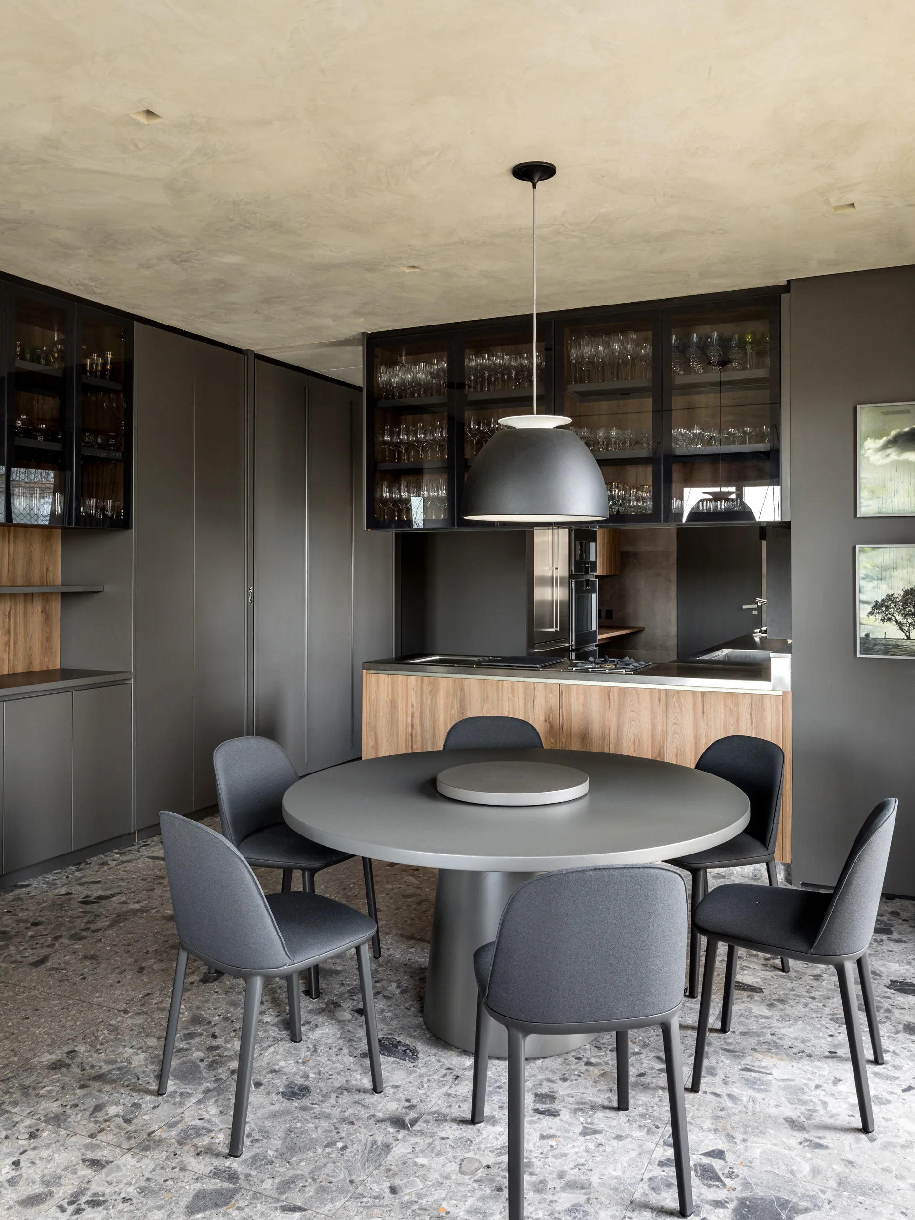 A round gray dining table and matching chairs sit on a speckled terrazzo floor beneath a dome pendant light in a charcoal and wood kitchen.