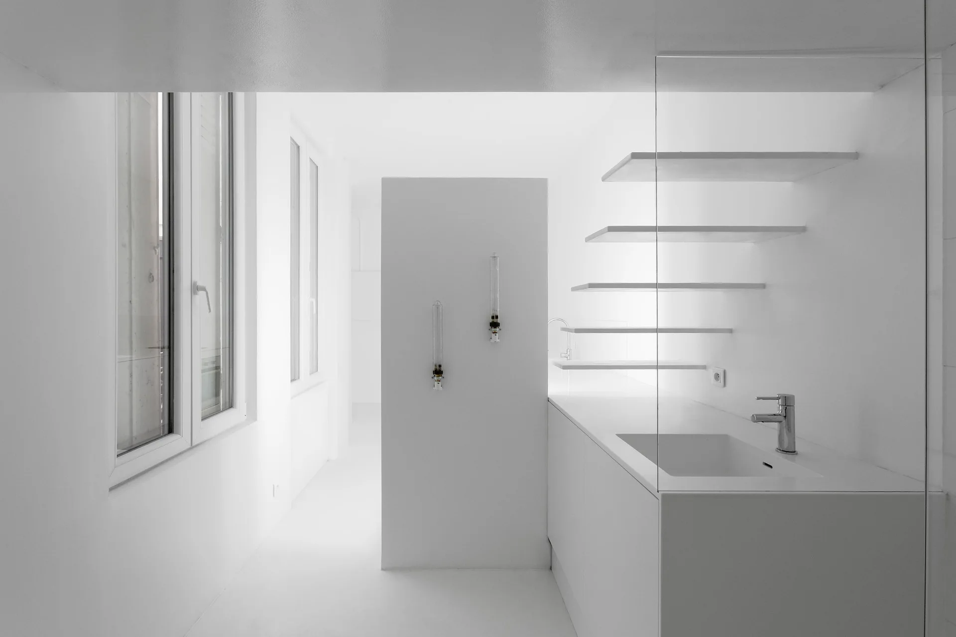 A stark minimalist white bathroom features a matte white vanity, floating shelves, a glass partition, and tall vertical windows.