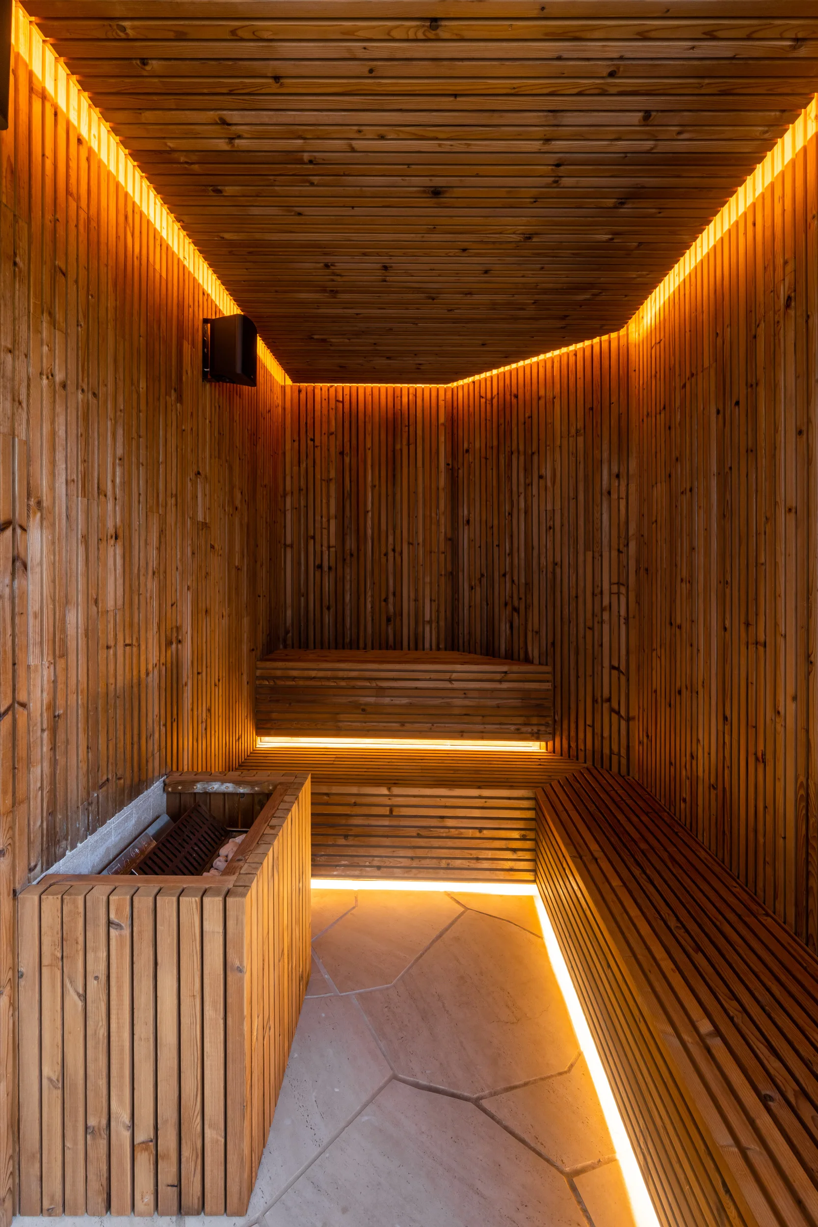 A modern sauna features vertical slatted wood walls, tiered wooden benches illuminated by integrated warm yellow lighting, and a stone tile floor.
