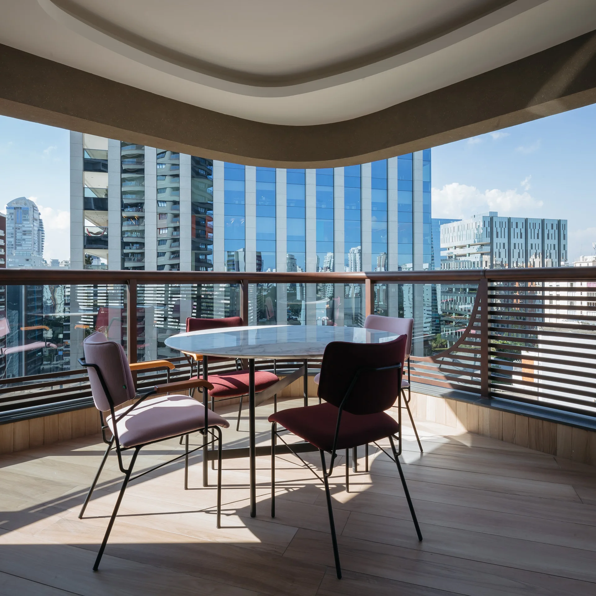 A round marble dining table with maroon and pink chairs sits on a wood-floored high-rise balcony overlooking the city skyline.