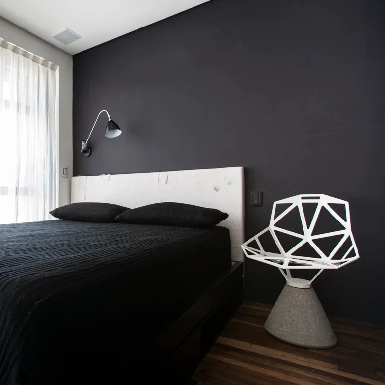 A modern bedroom features a black bed with a white headboard set against a matte black wall next to a white geometric sculptural chair.