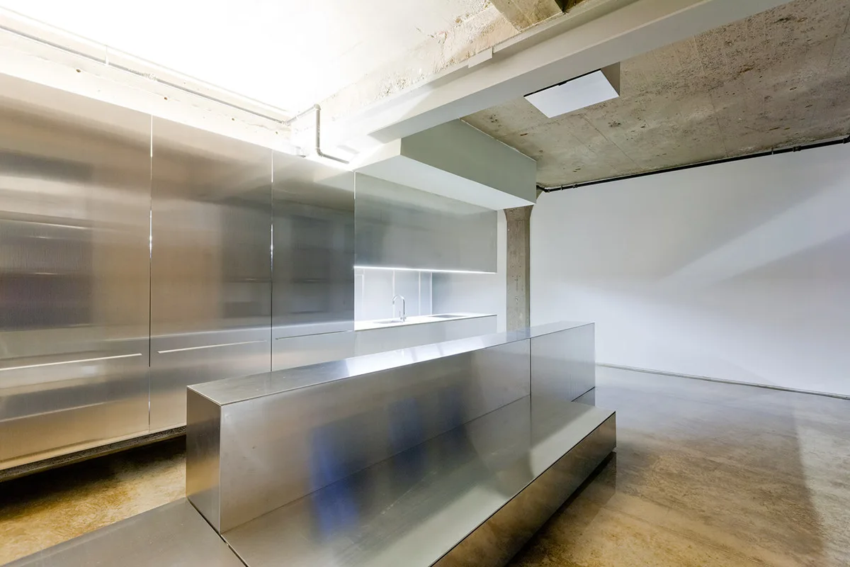 A minimalist kitchen features tall stainless steel cabinets, a tiered metal island, and an exposed raw concrete ceiling.