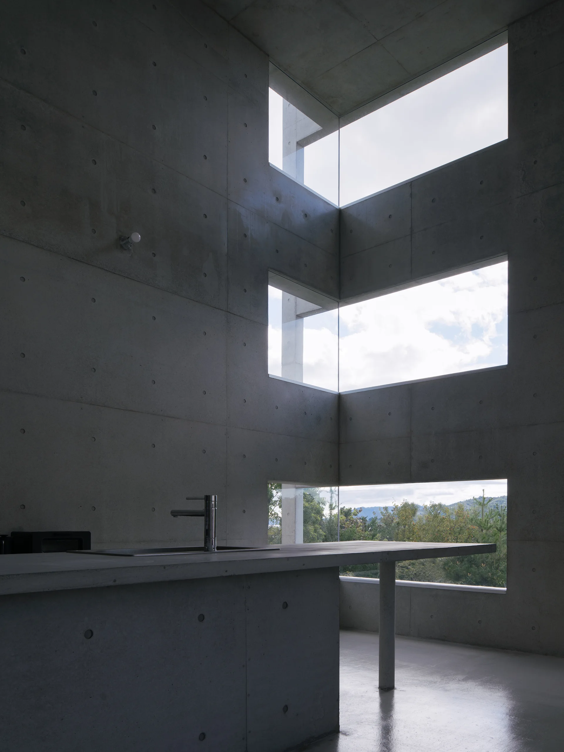 A gray concrete kitchen featuring a monolithic island with a faucet beneath three stacked corner windows that frame the sky and green trees.