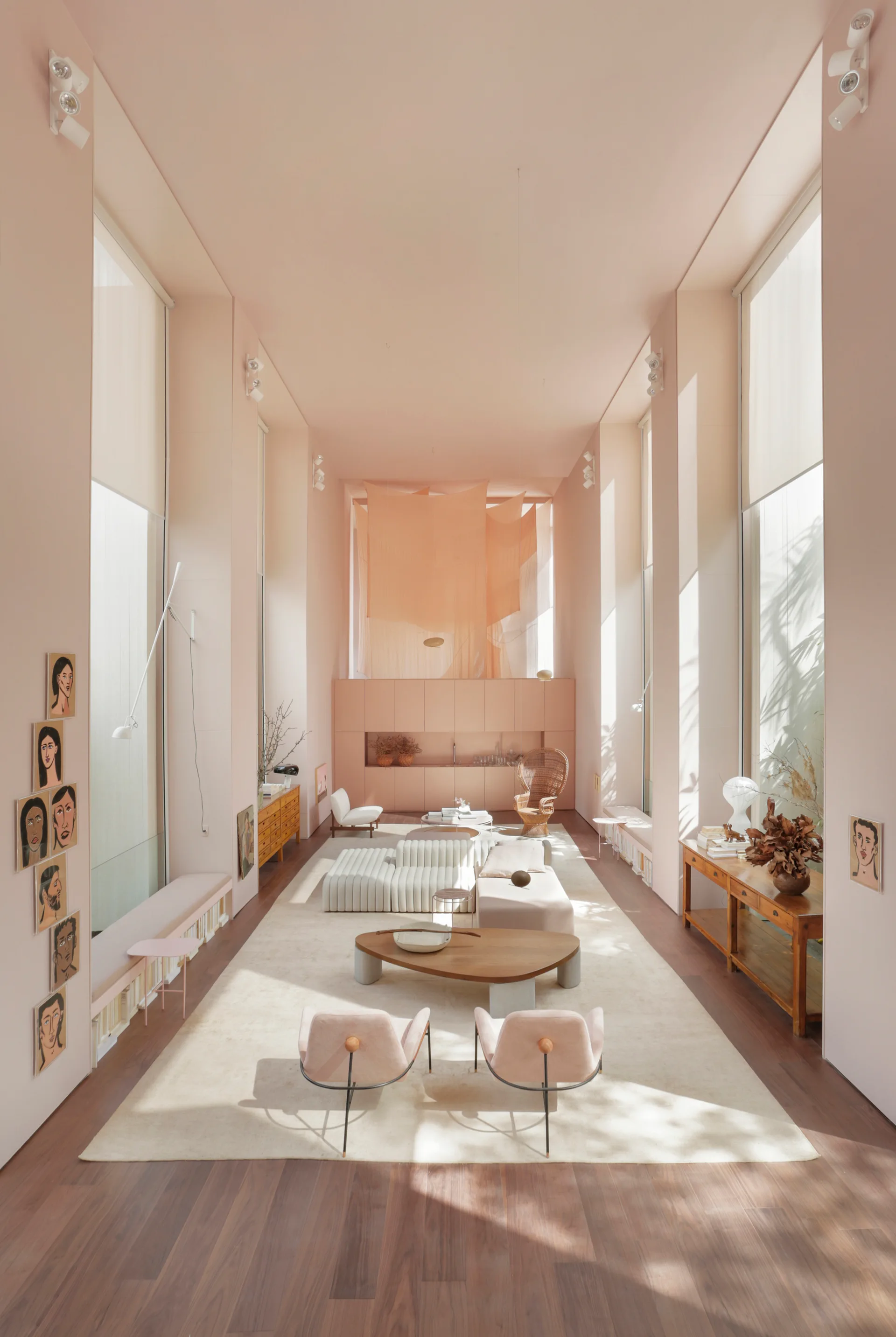 A tall monochromatic pink living room features a modular white sofa, wooden coffee tables, and built-in benches under high windows on wood floors.