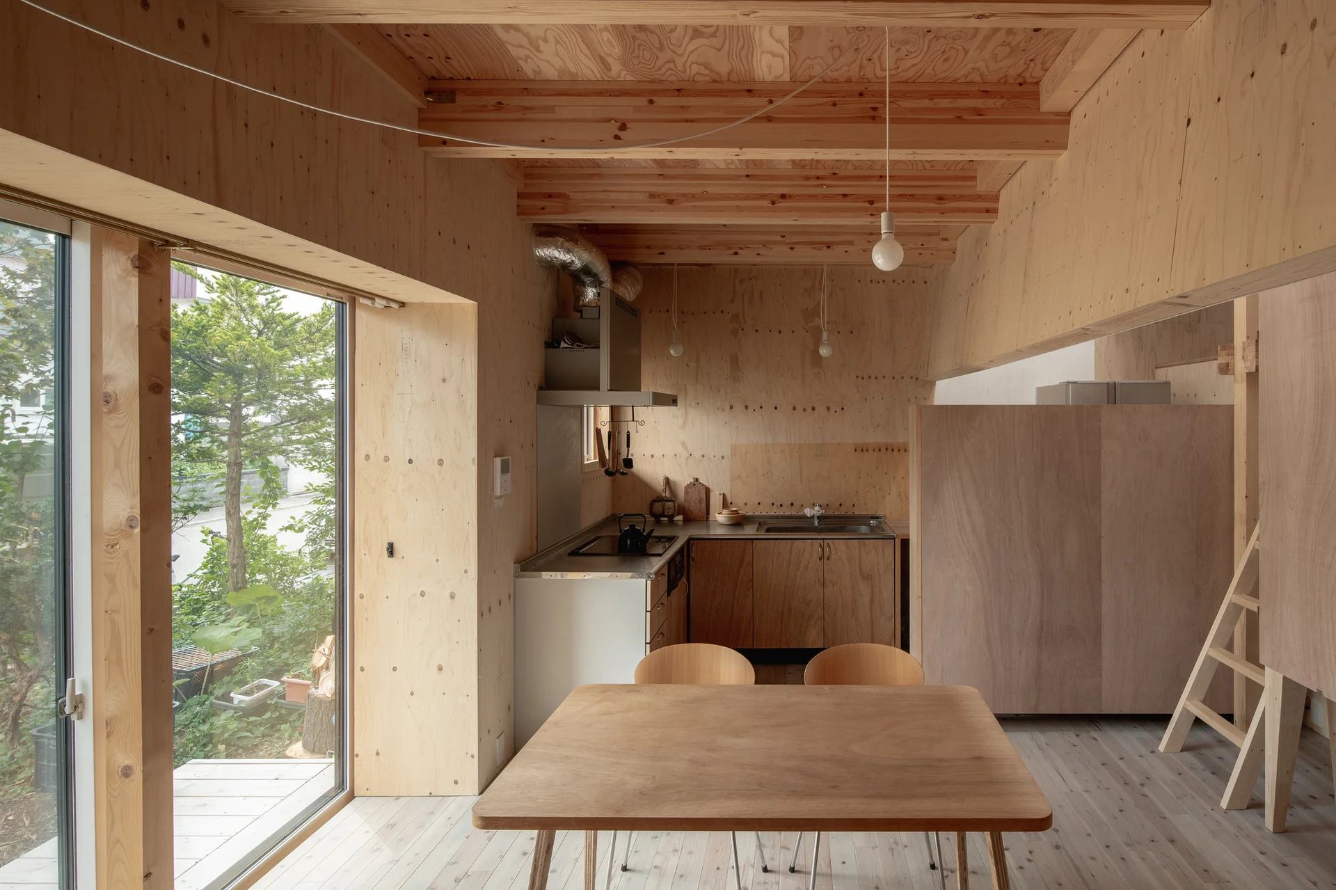 A minimalist kitchen and dining space featuring raw plywood walls, wooden ceiling beams, a simple wooden table, and a sliding glass door.