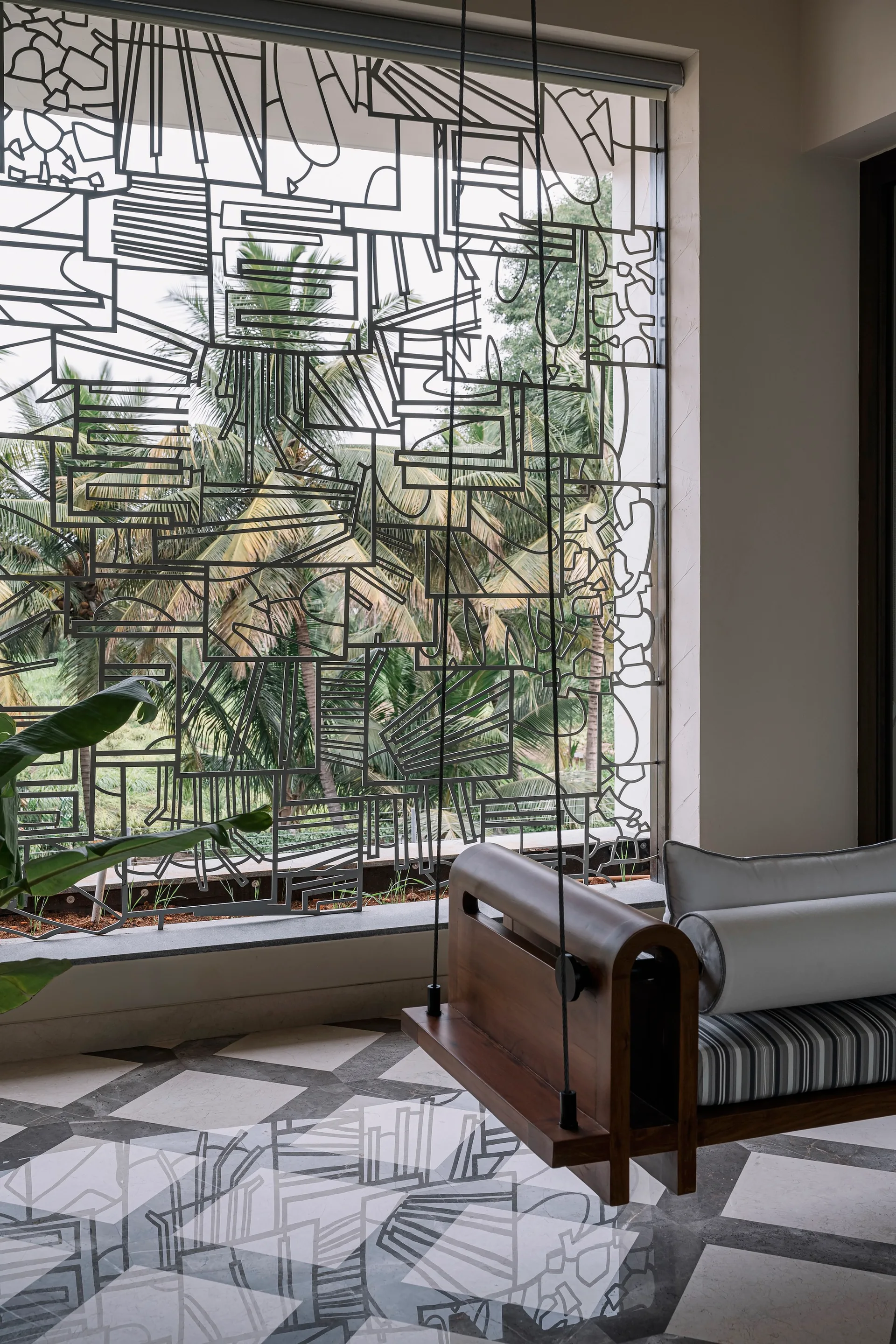 A dark wood hanging swing seat with striped and white bolster cushions sits near a large window featuring an intricate black geometric grille.
