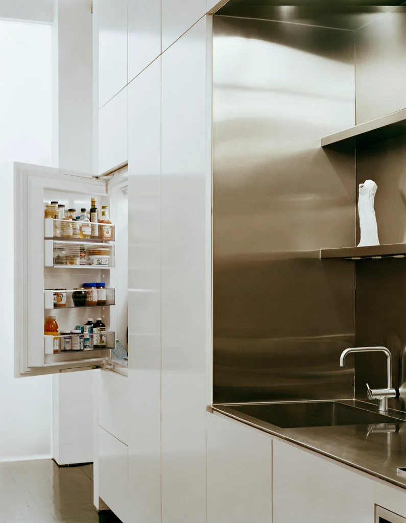 A minimalist kitchen features flat-panel white cabinetry, an open integrated refrigerator, and an inset stainless steel sink alcove.