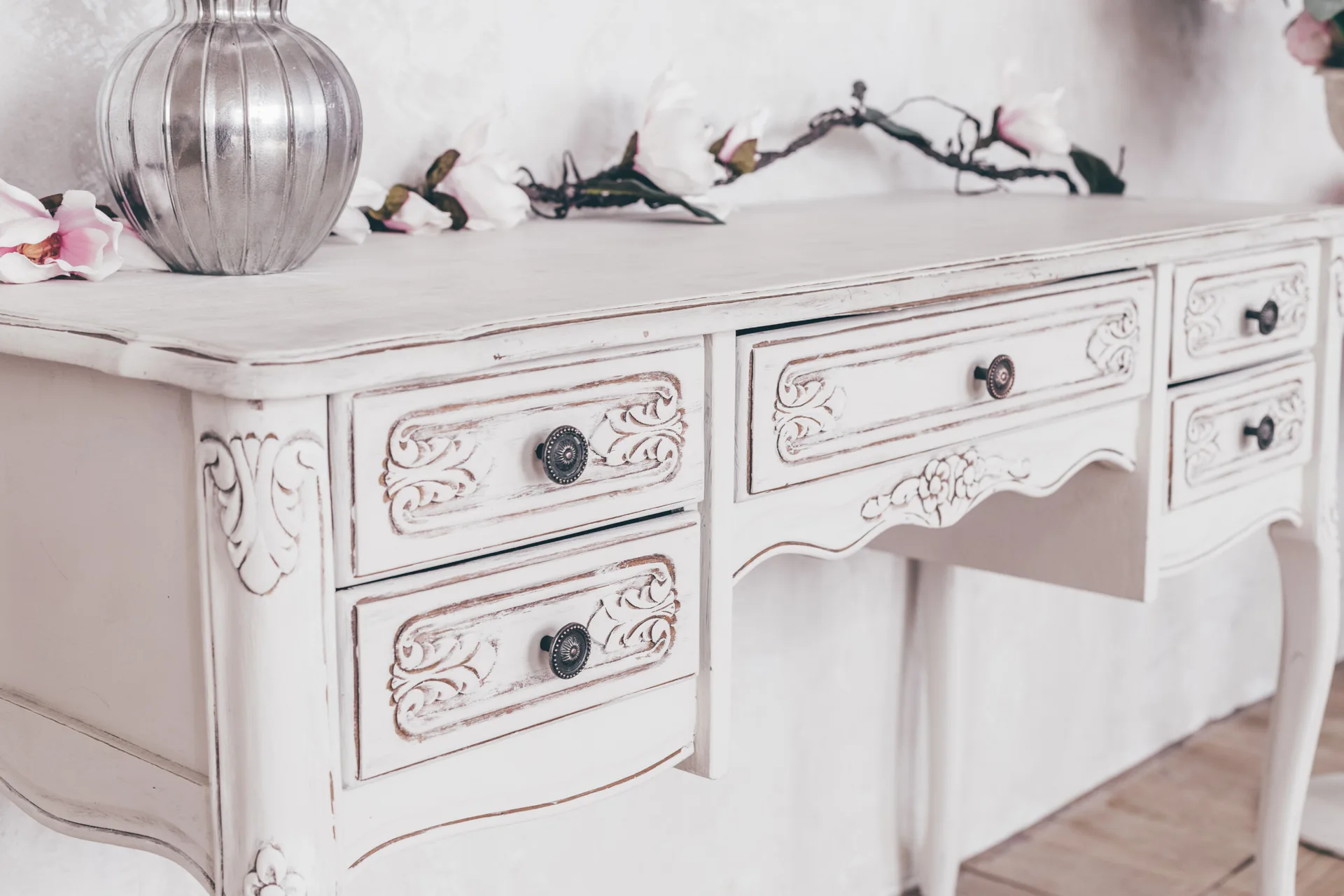 A distressed white wooden vanity desk with ornate carved drawers is decorated with a silver ribbed vase and pink flowers on top.