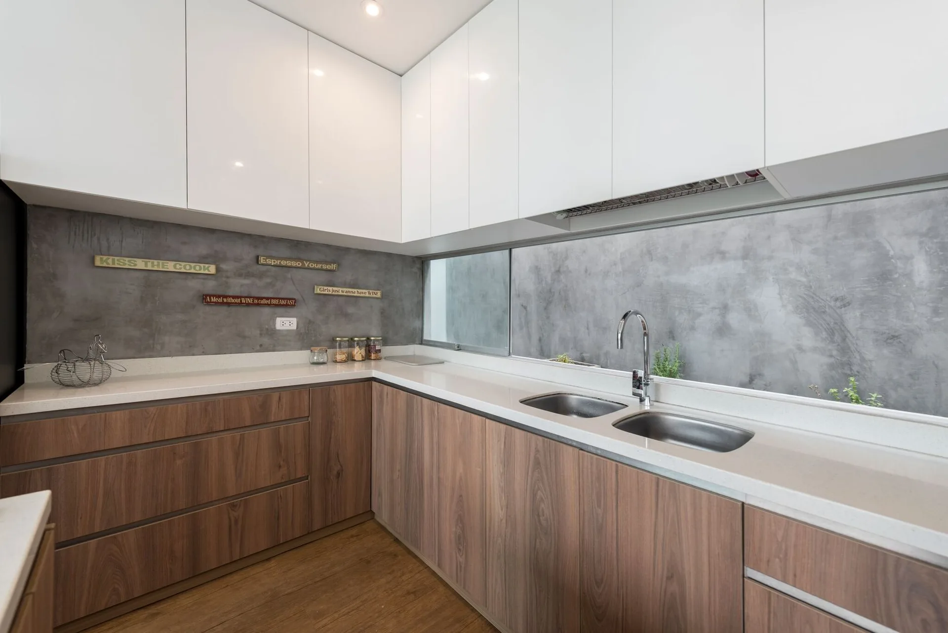 A modern kitchen with glossy white upper cabinets, wood-grained lower cabinets, and a grey cement-look backsplash with a horizontal window.