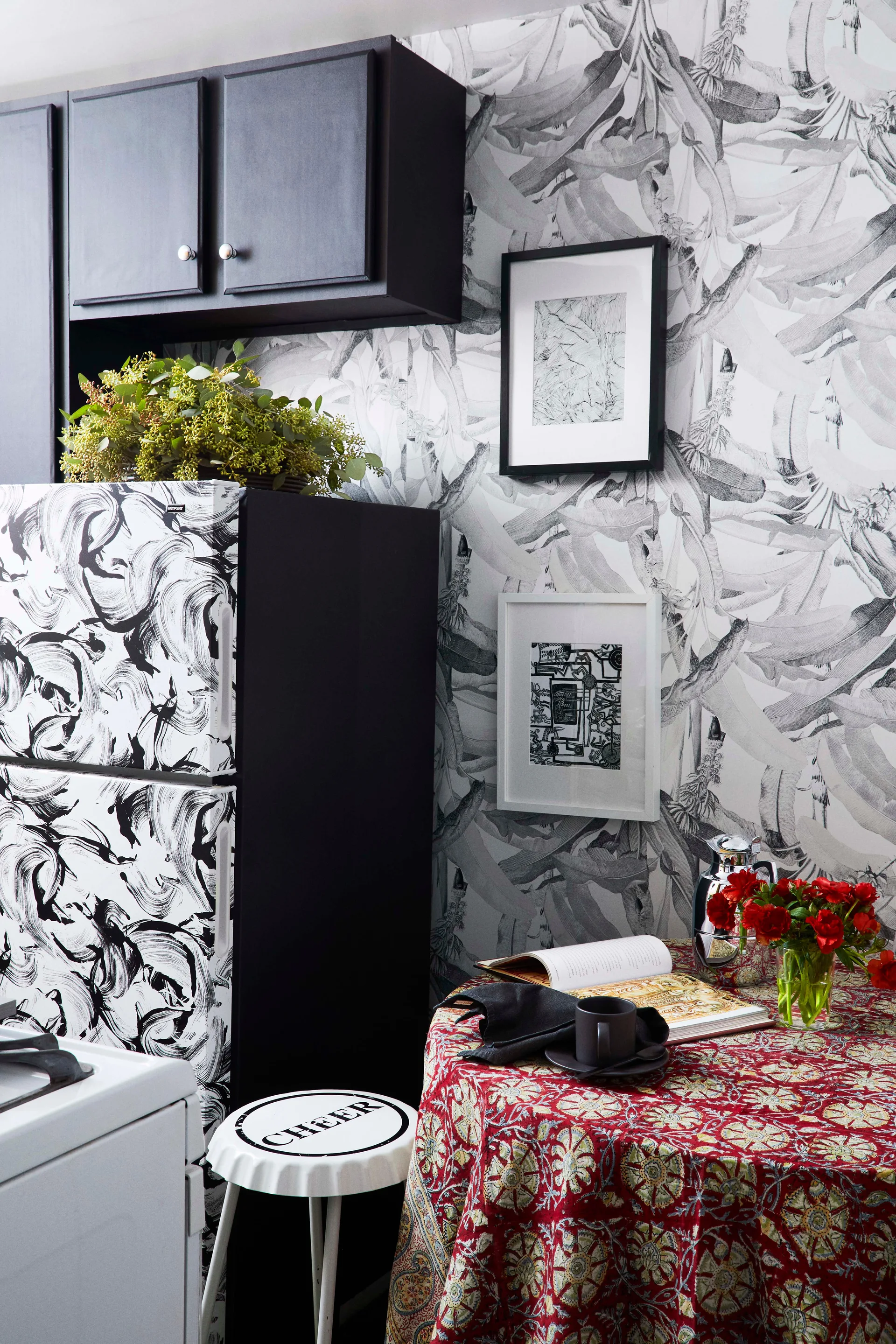 A kitchen nook features black and white botanical wallpaper, a patterned refrigerator, black upper cabinets, and a dining table with a red patterned tablecloth.