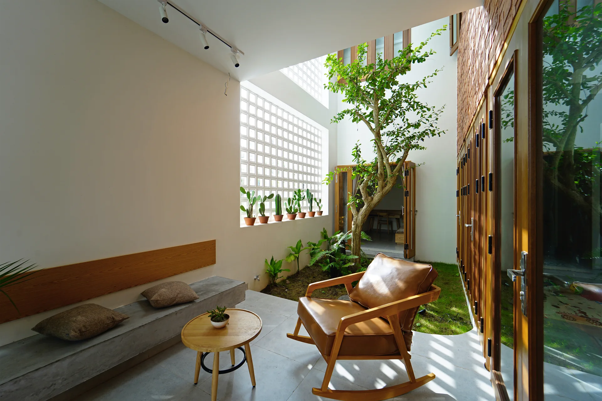 A light-filled indoor courtyard featuring a brown leather rocking chair, a concrete bench, and an integrated garden with a growing tree.