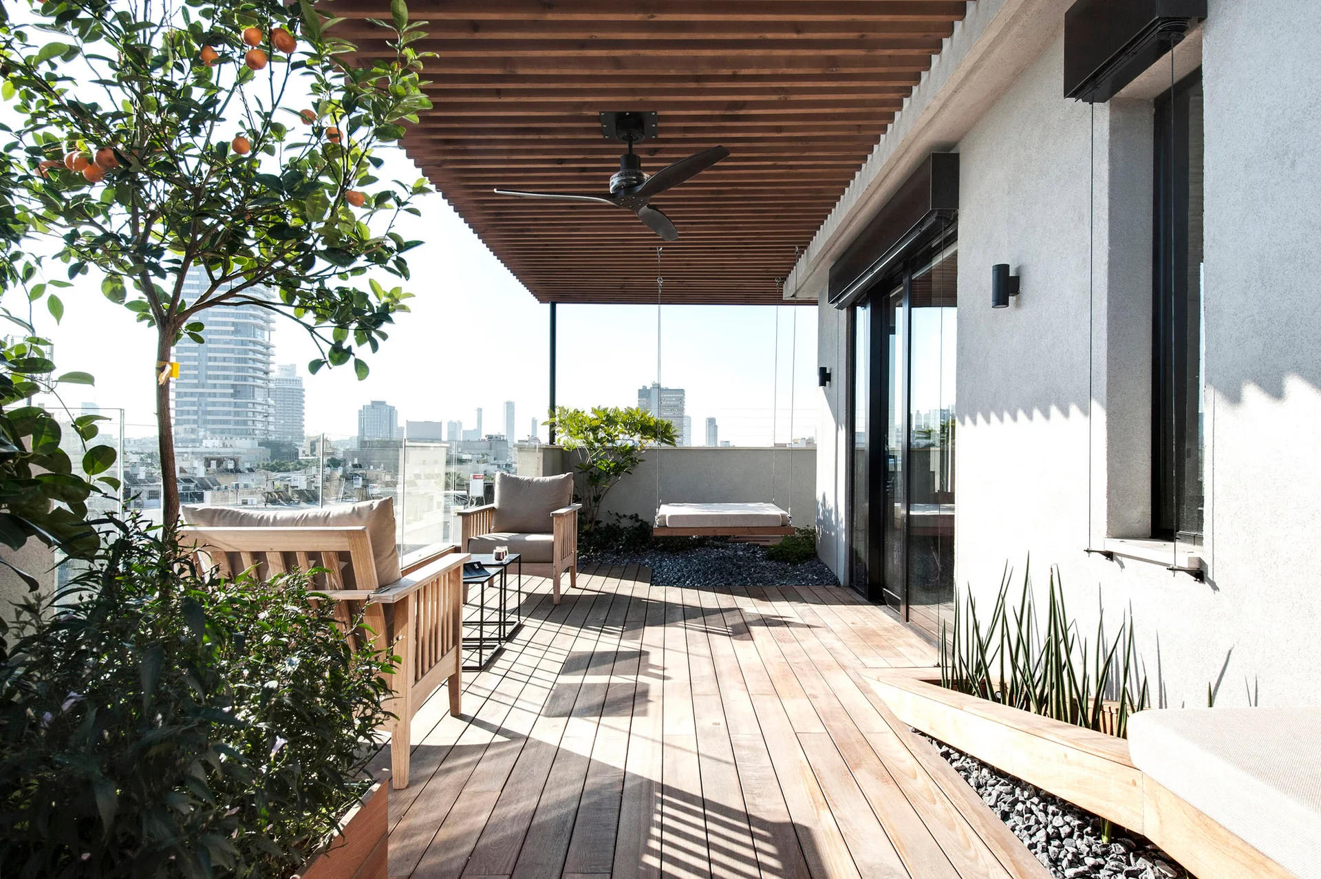 A modern rooftop terrace features wood deck flooring, a slatted timber ceiling with a black fan, wooden armchairs, and a hanging daybed.