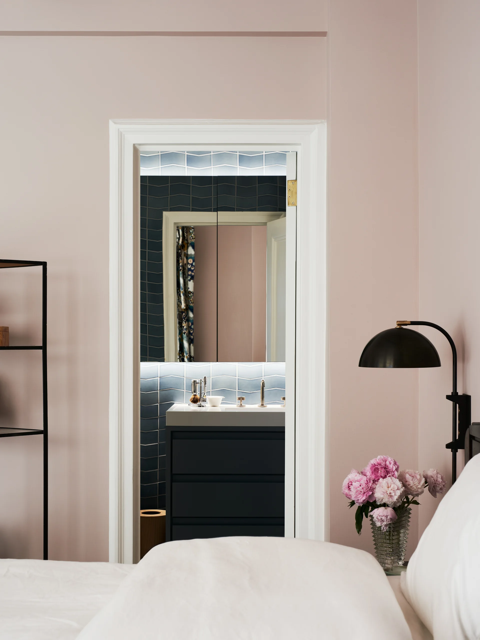A bedroom with dusty pink walls and a black wall lamp features a doorway looking into a bathroom with blue scalloped wall tiles.