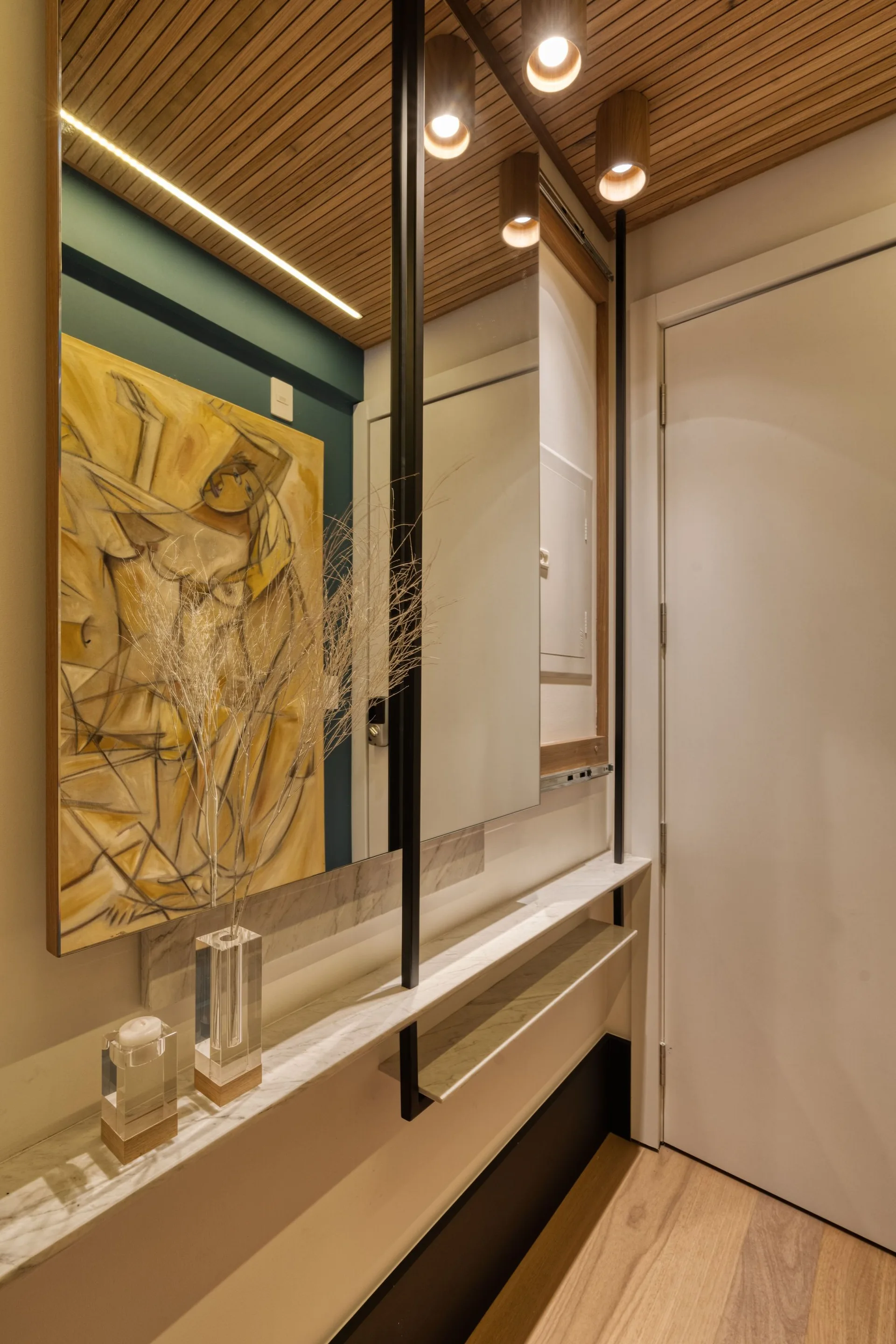 A modern entryway features a narrow marble shelf, a large mirror reflecting a teal wall and yellow artwork, and a wood slat ceiling with spotlight fixtures.