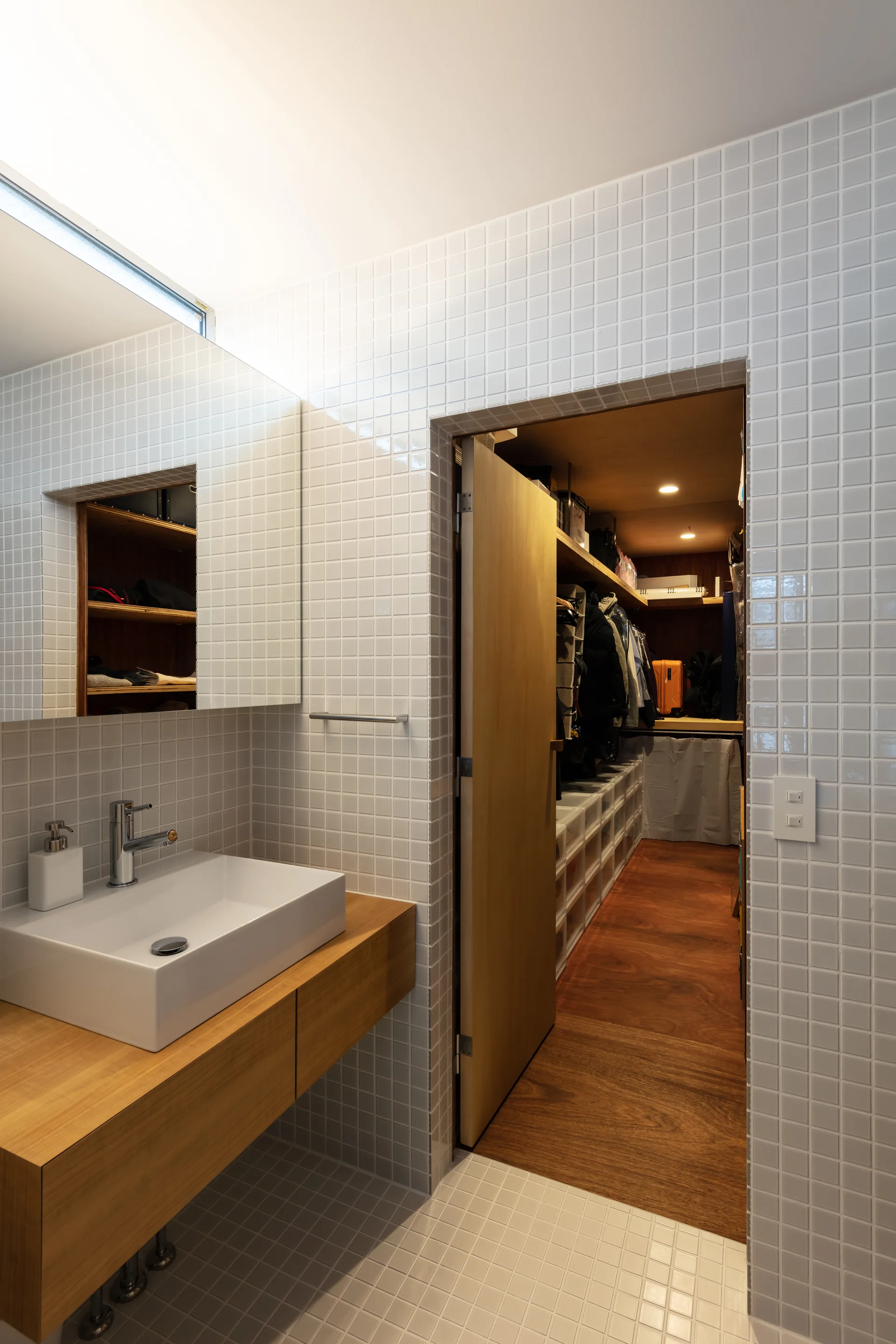 A modern bathroom features white grid tile walls and a floating wooden vanity next to an open doorway leading to a walk-in closet.