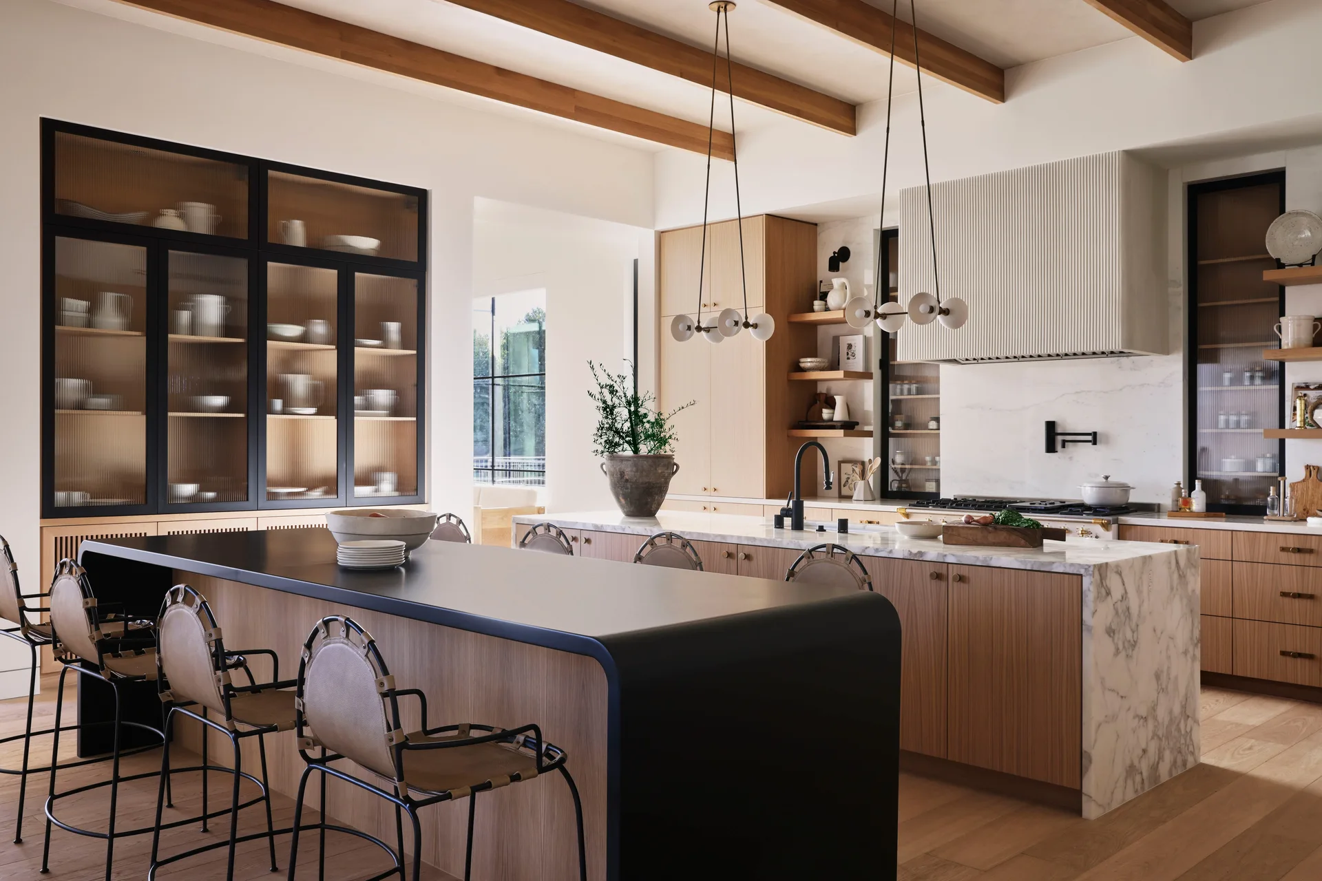 A contemporary kitchen features a double-tiered island with bar stools, light wood cabinets, exposed ceiling beams, and a white fluted range hood.