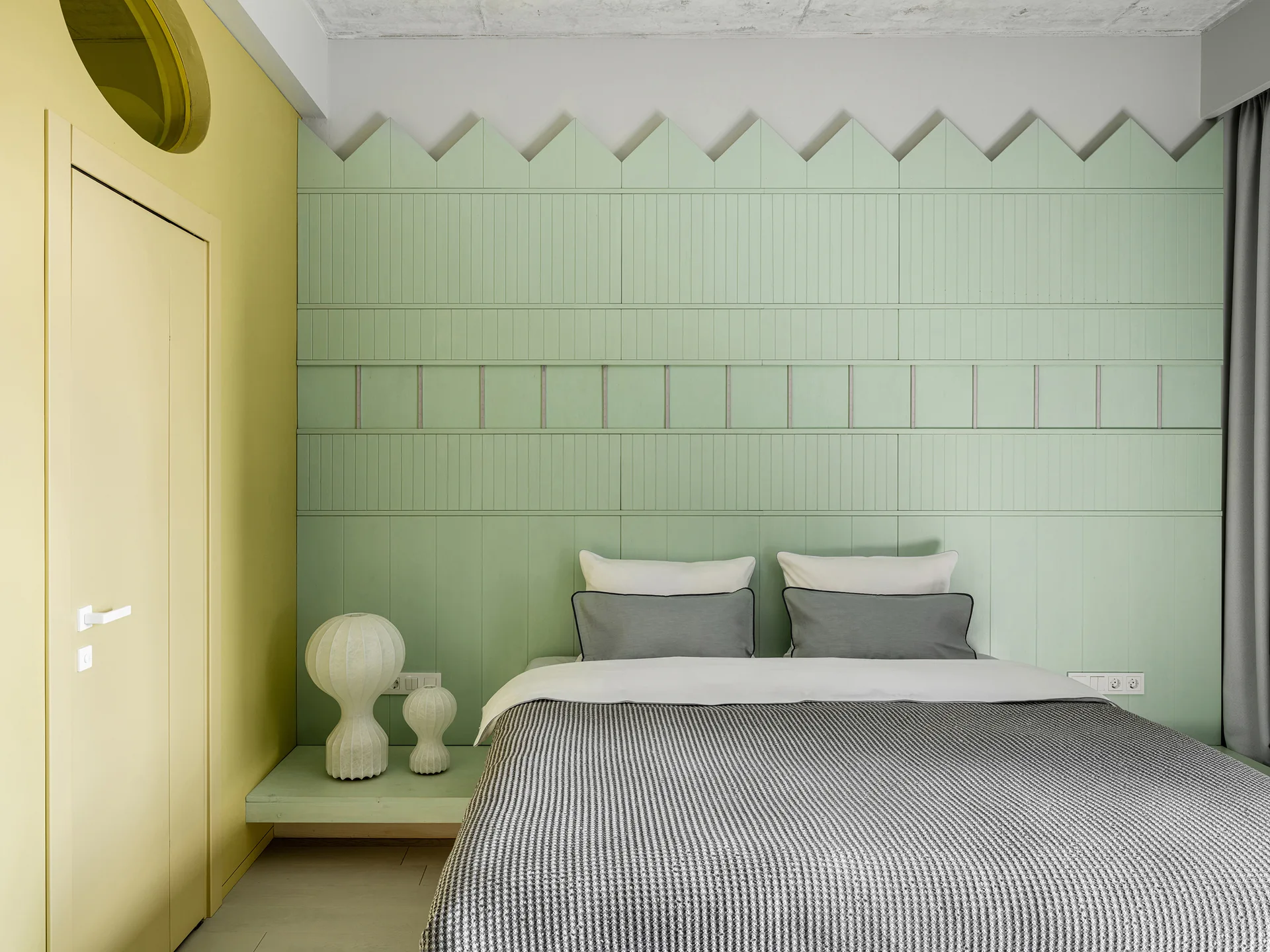A modern bedroom features a mint green picket-style wood-paneled wall behind a bed, a yellow accent wall with a door, and two sculptural bedside lamps.